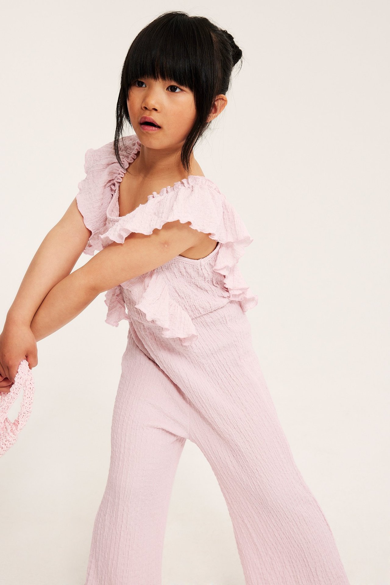 Widok z przodu: Dziecko w jasnoróżowym kombinezonie z dzianiny ze ściąganym pasem, falbankami i szerokimi, krótkimi nogawkami.