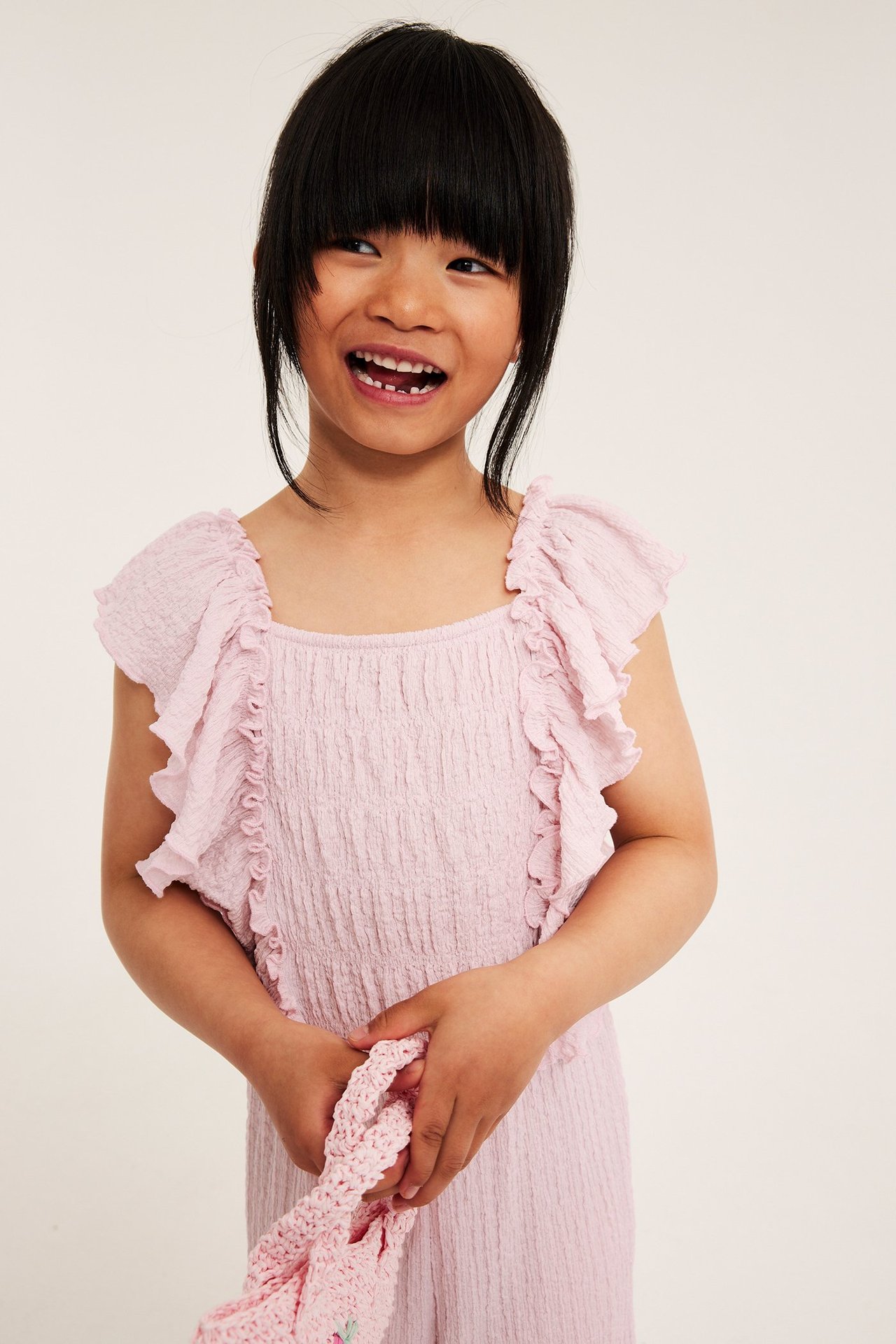 Dziecko w jasnoróżowym kombinezonie z falbankami na ramionach, trzyma różowy dziergany przedmiot.