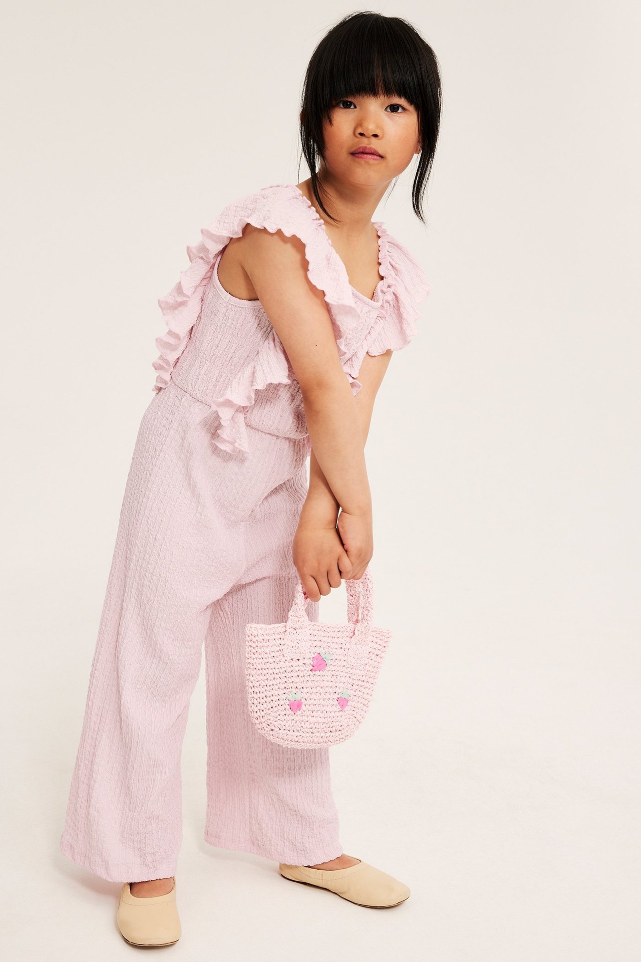 Edestä: Lapsi yllään vaaleanpunainen trikoohaalari olkalaukkufrilloilla, pitäen vaaleanpunaista mansikkapussukkaa.