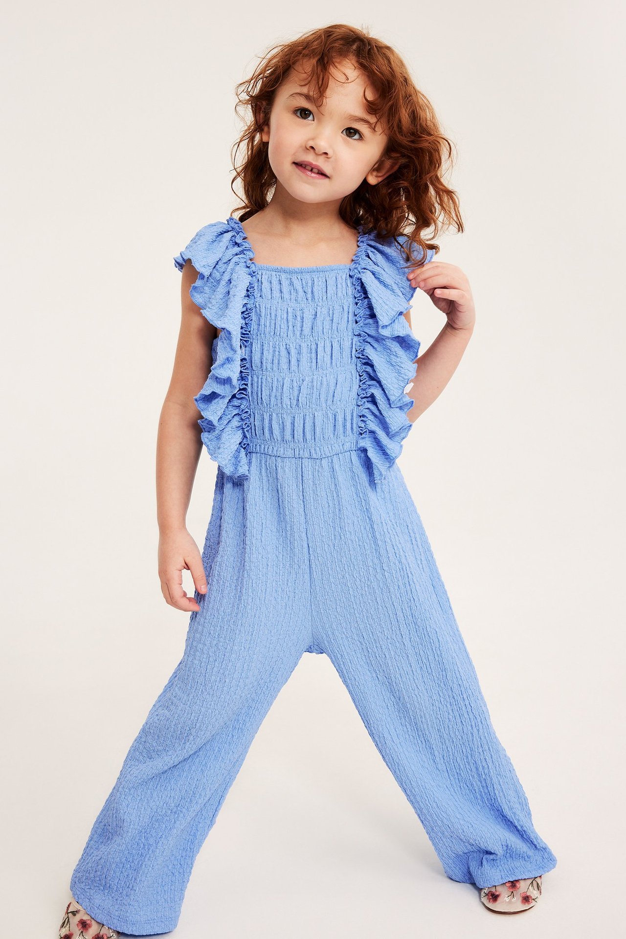 Widok z przodu: dziecko w niebieskim dżersejowym kombinezonie z falbanami, smockiem, szerokimi krótkimi nogawkami i butami.
