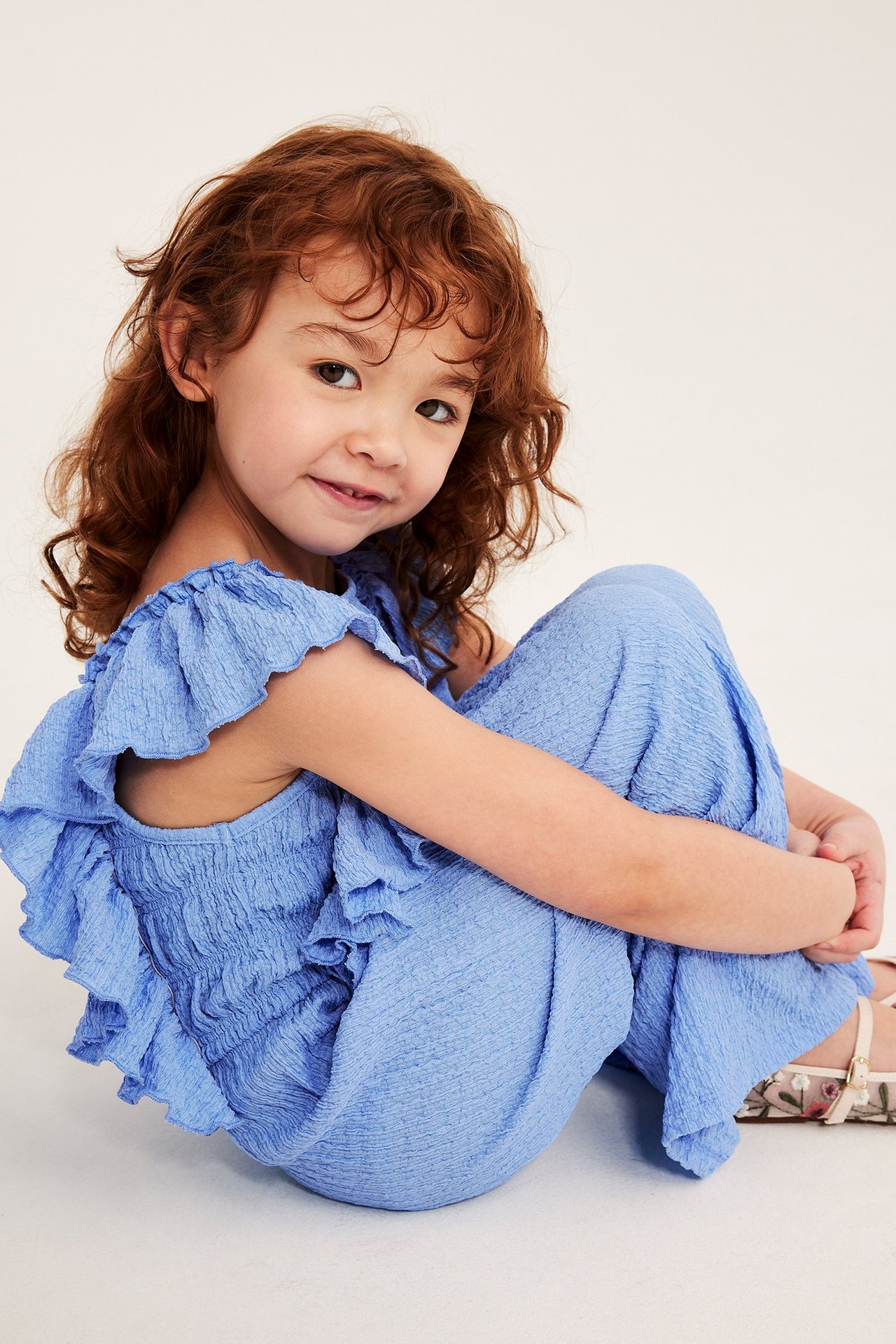 Edestä kuvattu lapsi sinisessä jerseyhaalariasussa, jossa on smokkirypytys, olkapääfrilloja ja kukkakengät.