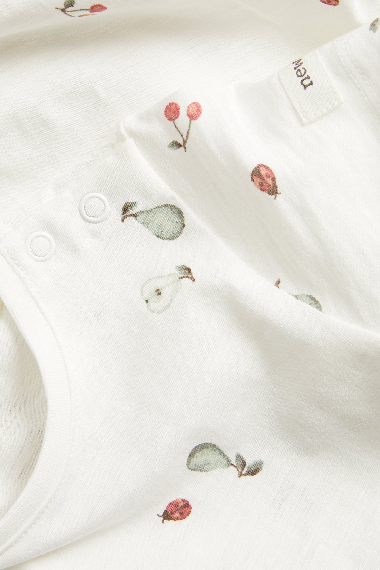 Lähikuva luonnonvalkoisesta vauvan body-paidasta, jossa on päärynä-, kirsikka- ja leppäkerttukuviot, painonapeilla.