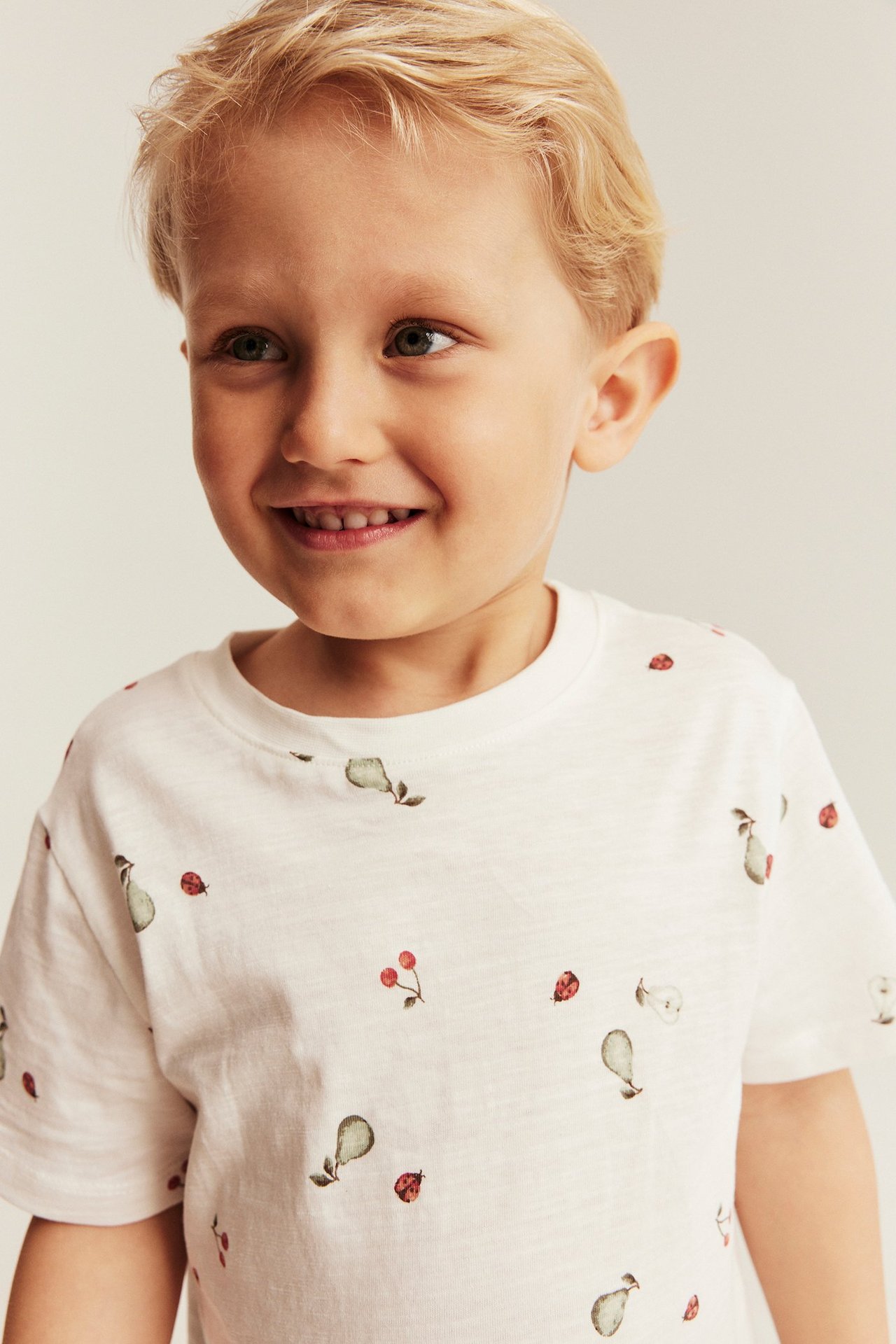 Edestä, hymyilevä poika offwhite-slubtrikoo-paidassa päärynä-, kirsikka- ja leppäkerttuprintillä.