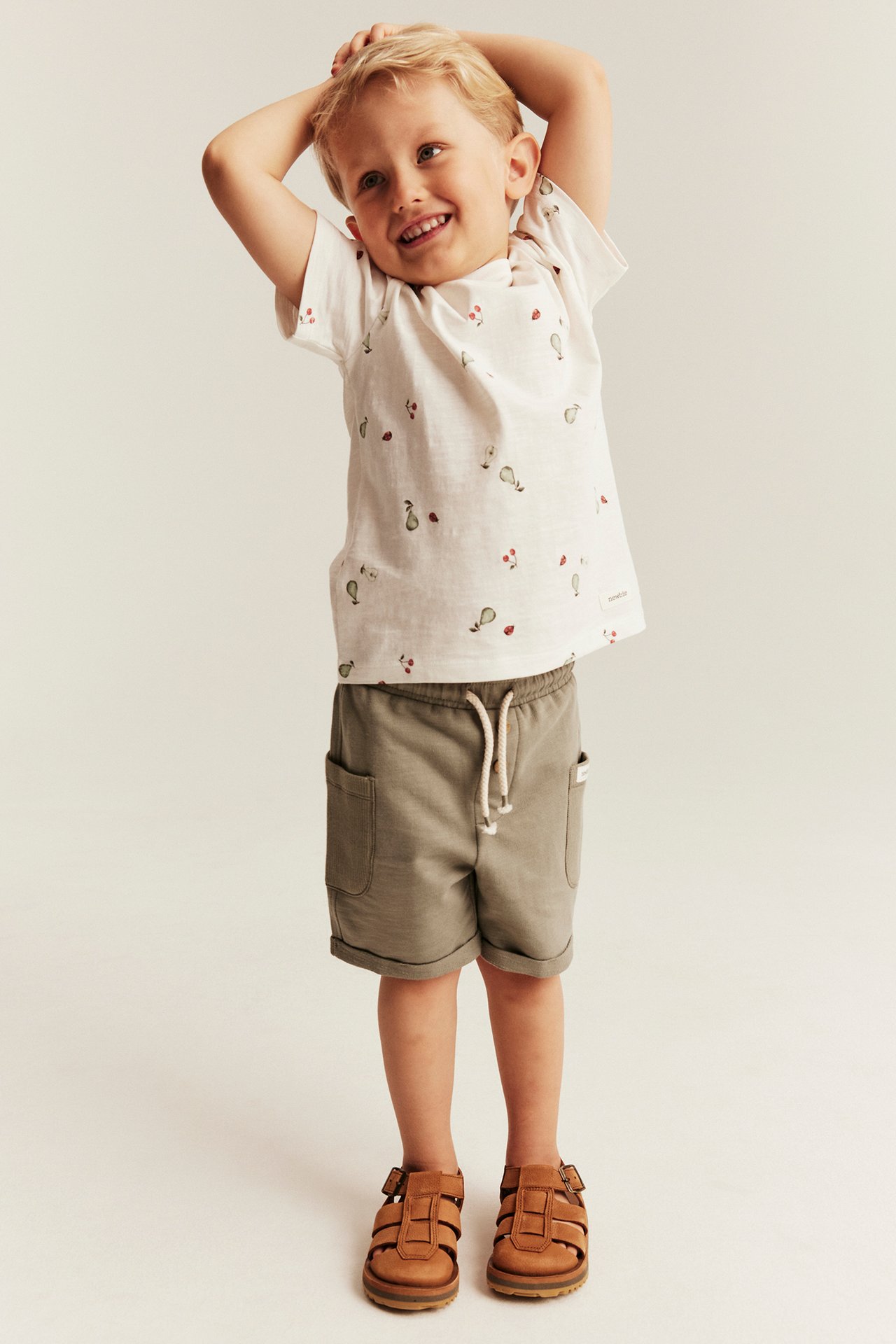 Näkymä edestä: lapsi luonnonvalkoisessa T-paidassa hedelmäkuviolla, khakishortseissa ja ruskeissa sandaaleissa.