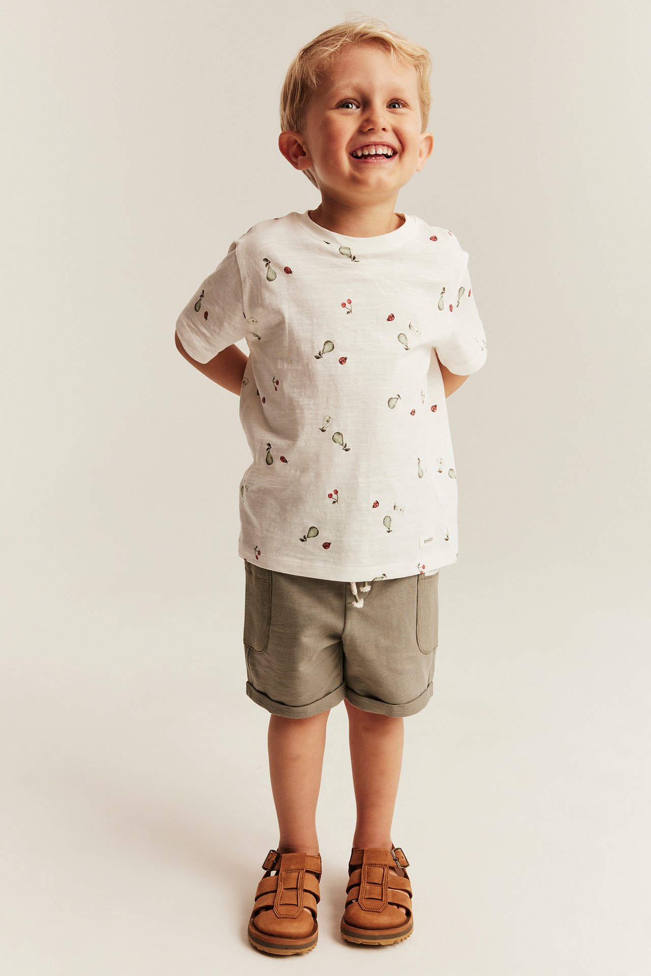 Hymyilevä lapsi offwhite t-paidassa päärynöillä, kirsikoilla, leppäkertuilla, khakishortseissa ja ruskeissa sandaaleissa.