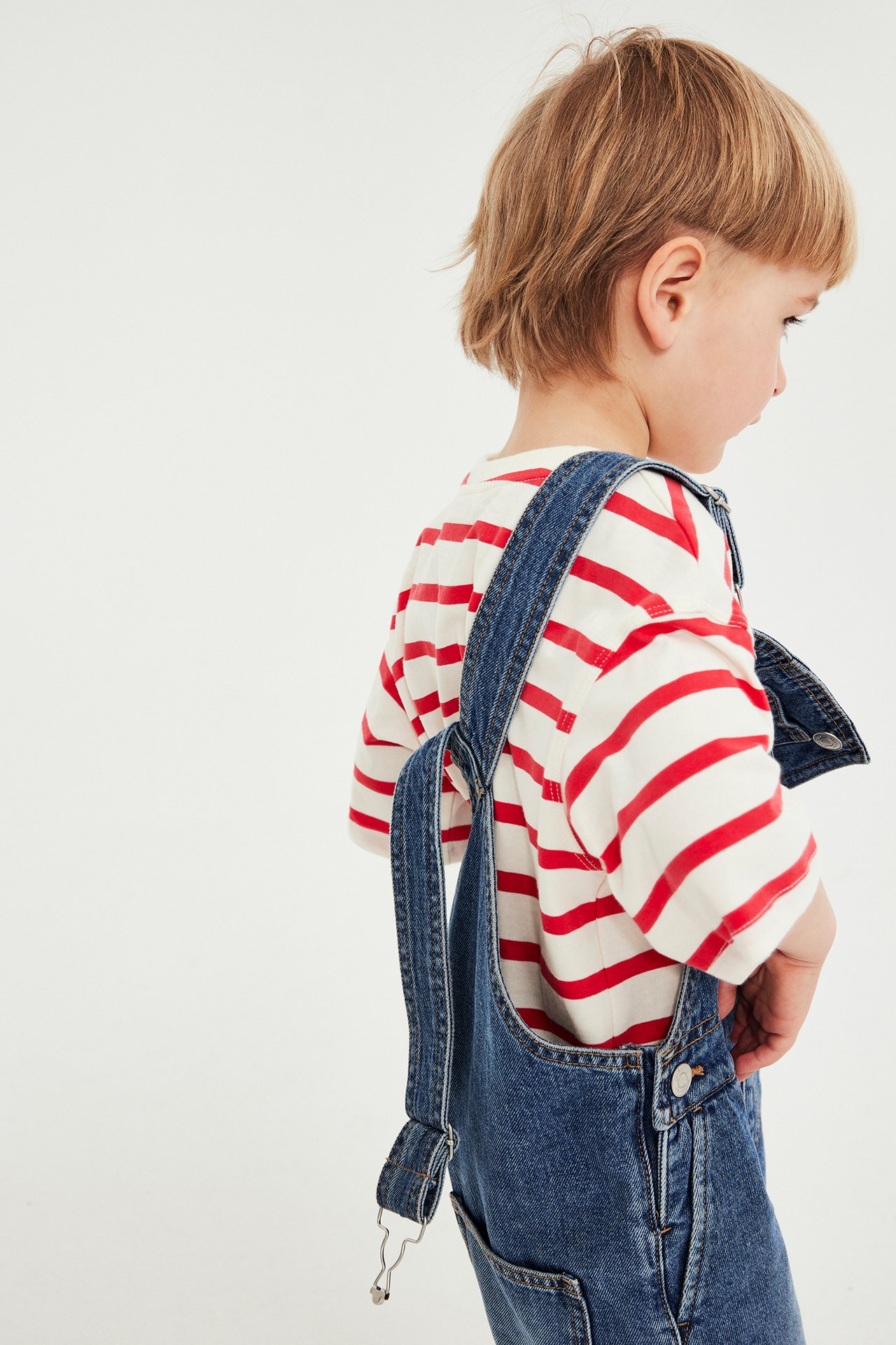 Sidovy av ett barn i vit- och rödrandig kortärmad bomullst-shirt under blå denimoveraller.