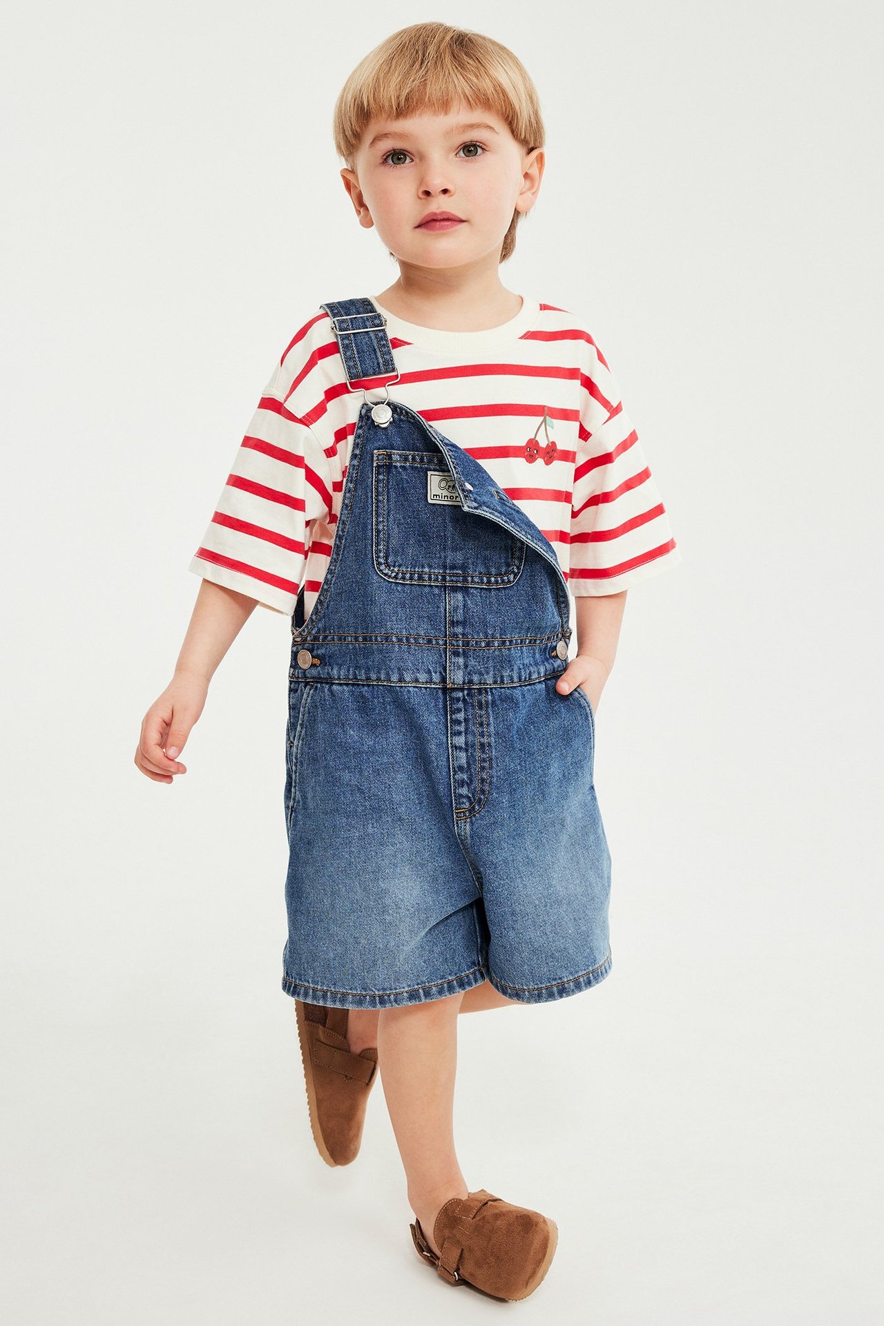 Widok z przodu: dziecko w czerwono-białej koszulce w paski z haftem wiśni i niebieskich dżinsowych ogrodniczkach.
