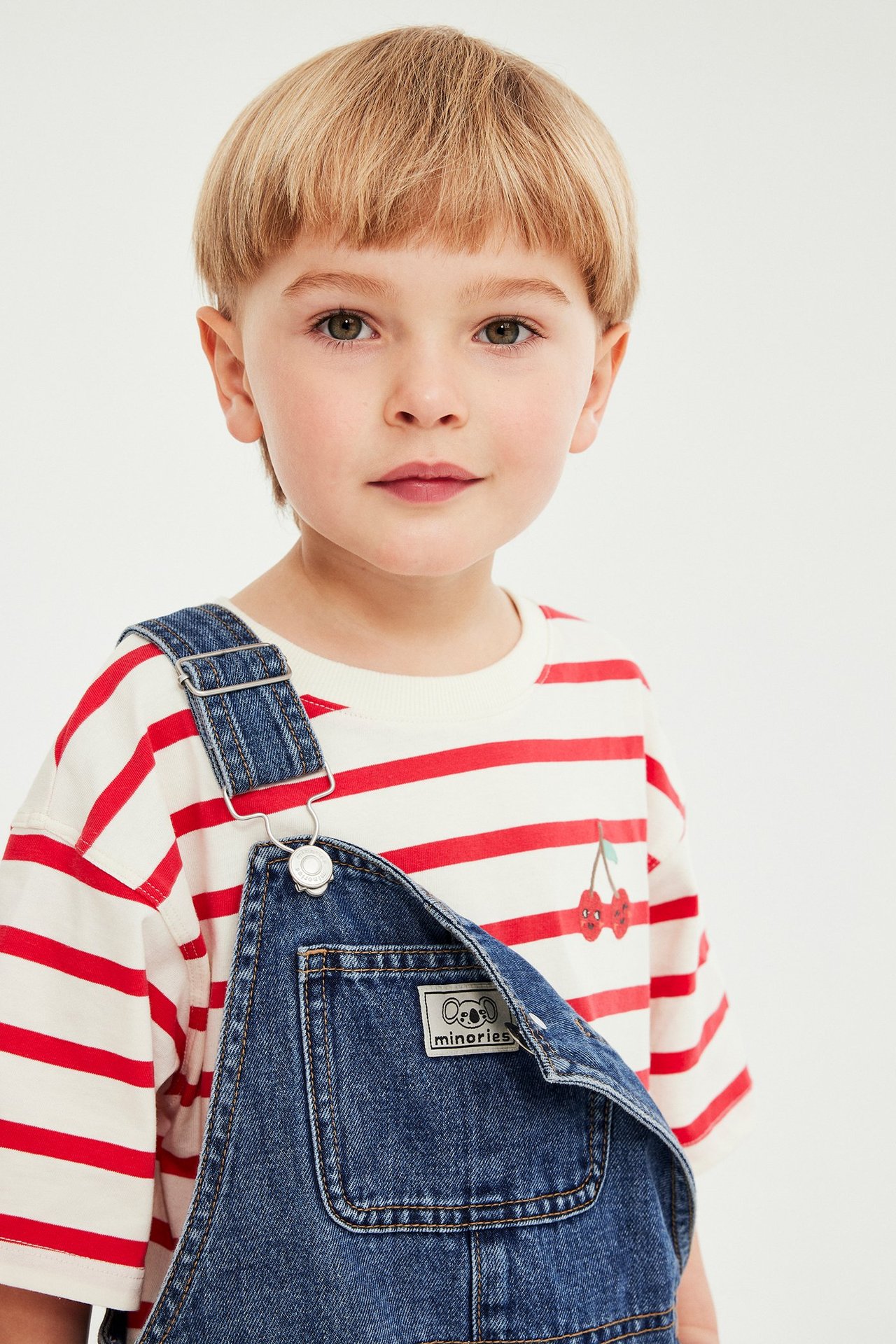 Frontvy: Ett barn i vit- och rödrandig t-shirt med körsbärsbrodyr, under blå denimhängselbyxor.