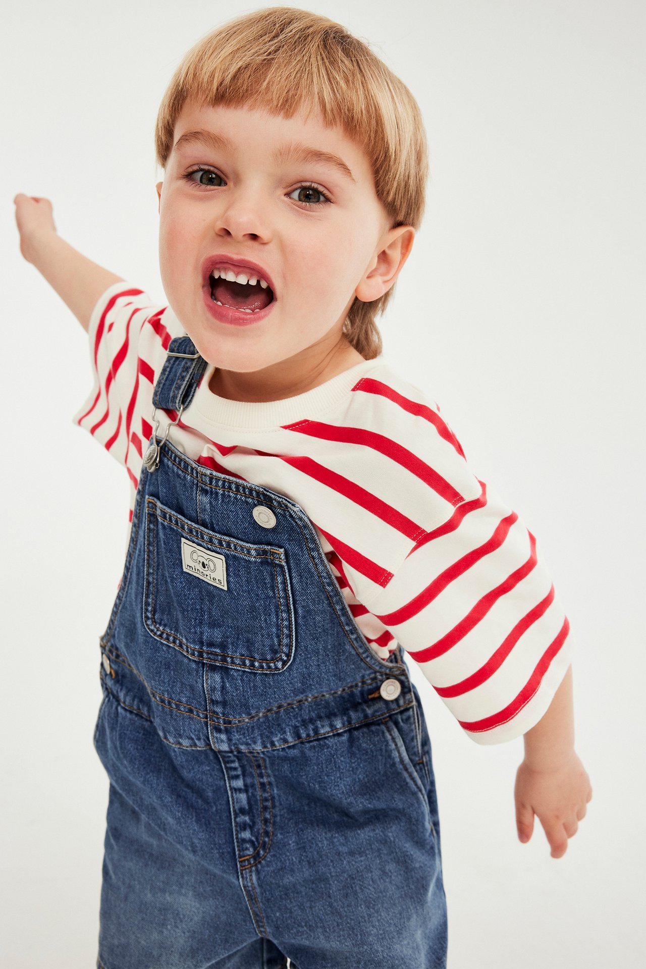 Frontvy av ett barn i en krämfärgad kortärmad t-shirt med röda ränder under blå jeanshängselbyxor.