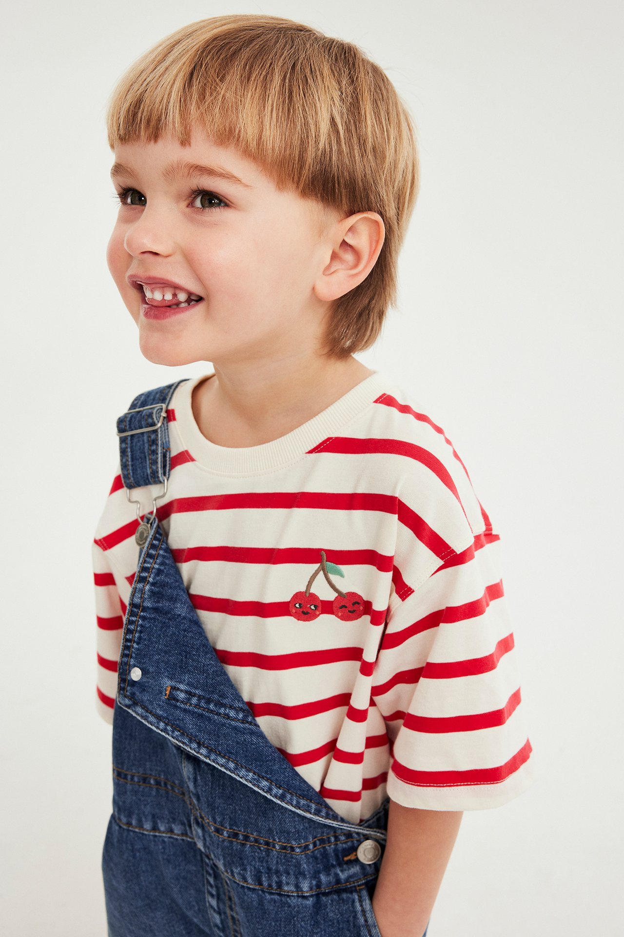 Przód: Dziecko w kremowym t-shircie w czerwone paski z haftem wiśni, pod dżinsowymi ogrodniczkami.