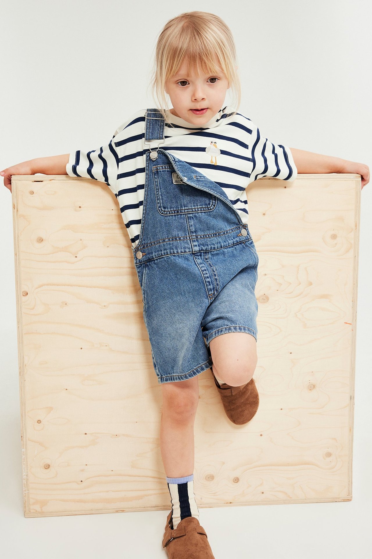 Framifrån: Barn i vit-blårandig bomulls-t-shirt med ankbrodyr, blå denimshorts-overall och bruna skor.