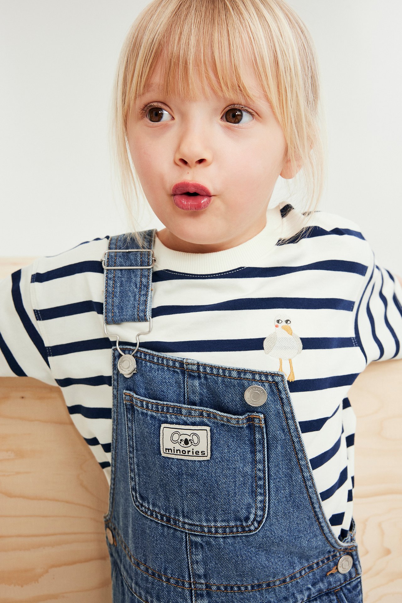 Närbild: Barn bär vit-marinblårandig t-shirt med broderad mås, under blå denimoveraller.