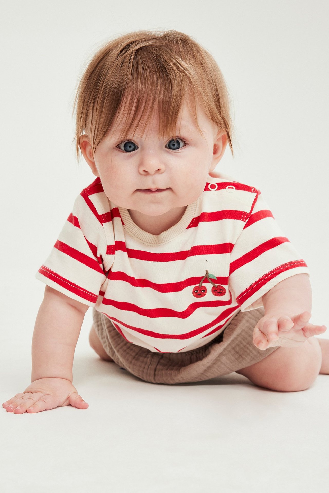 Krypande baby i röd- och vitrandig body med körsbärsbrodyr och ljusbruna shorts, framifrån.