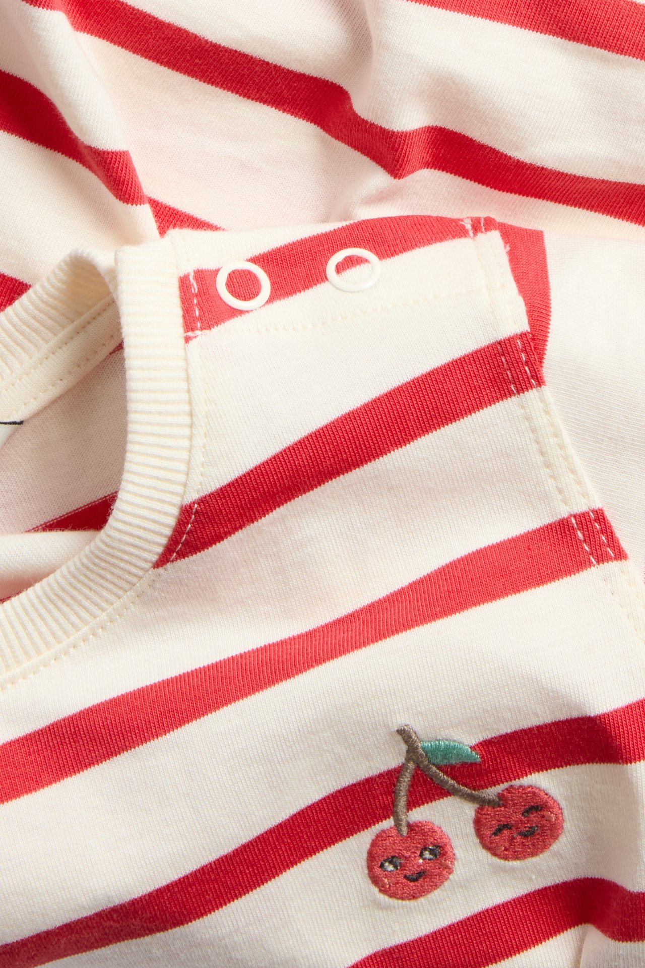 Lähikuva punavalkoraidallisesta puuvillavauvabody-paidasta olkapään neppareilla ja kirsikkakirjonnalla.