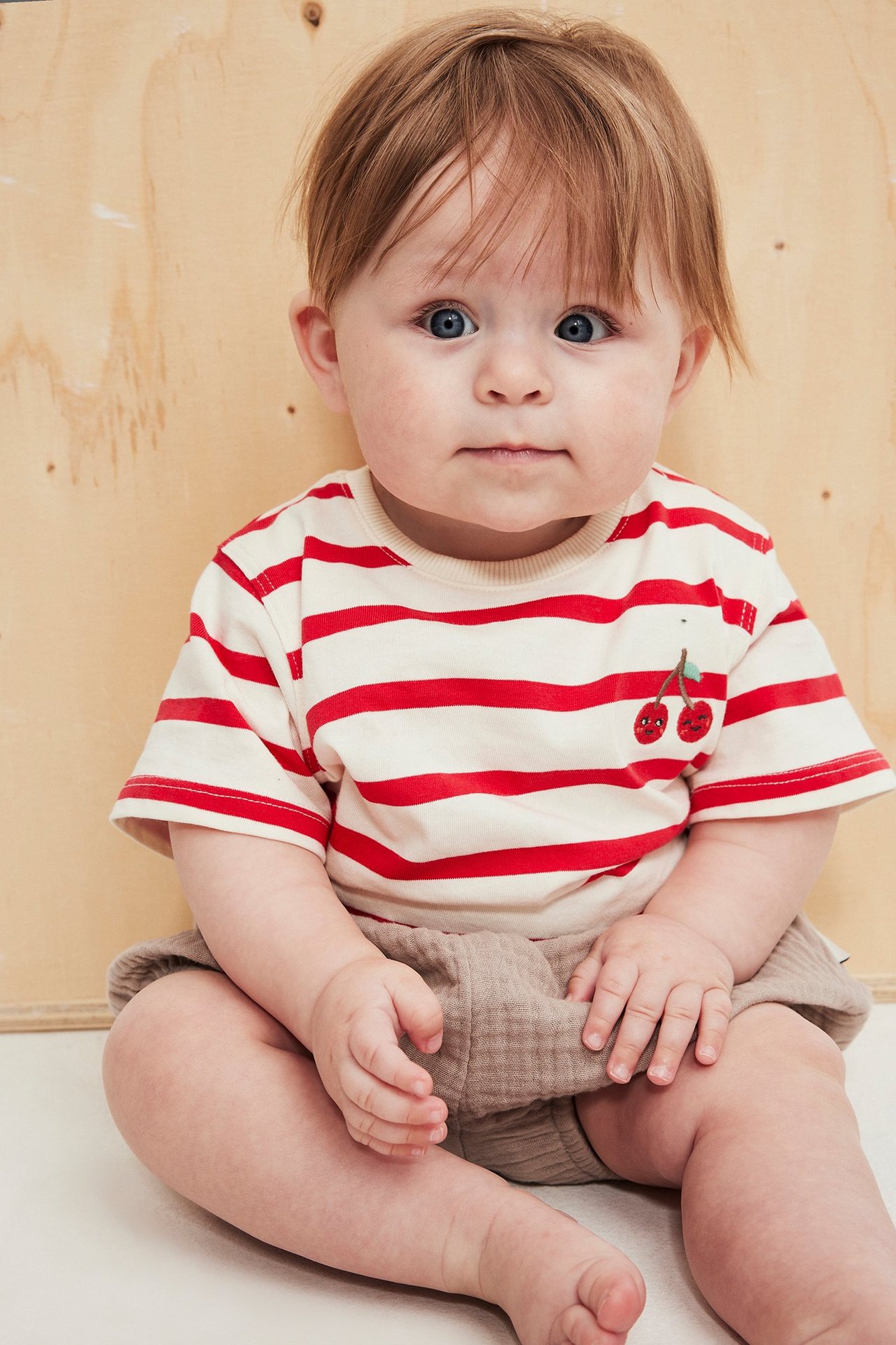 Baby i randig röd/crémefärgad kortärmad body med körsbärsbrodyr och beiga shorts, framifrån.