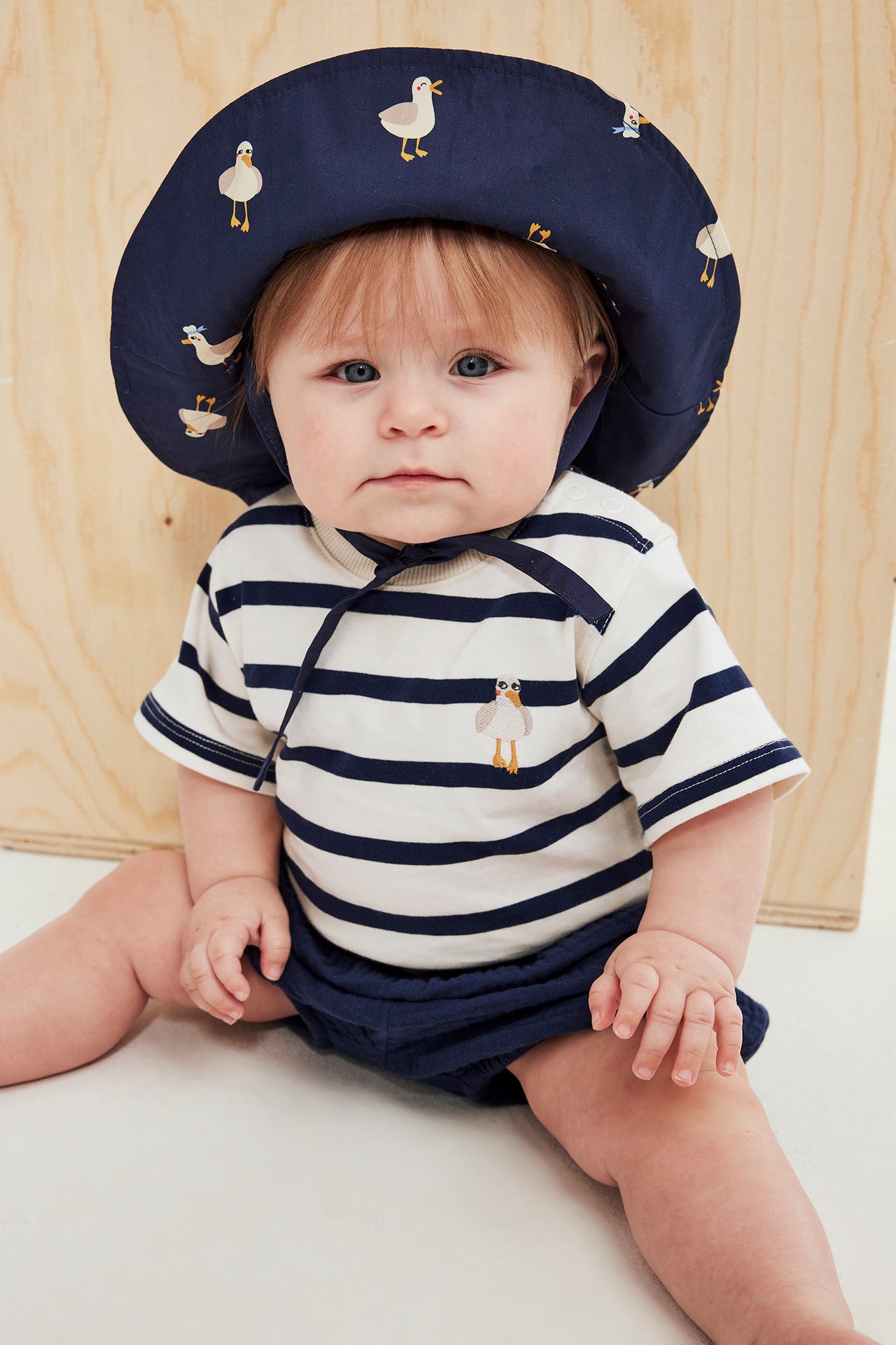 Frontbilde av en baby i stripete body med måkebroderi, marineblå shorts og en marineblå mønstret hatt.