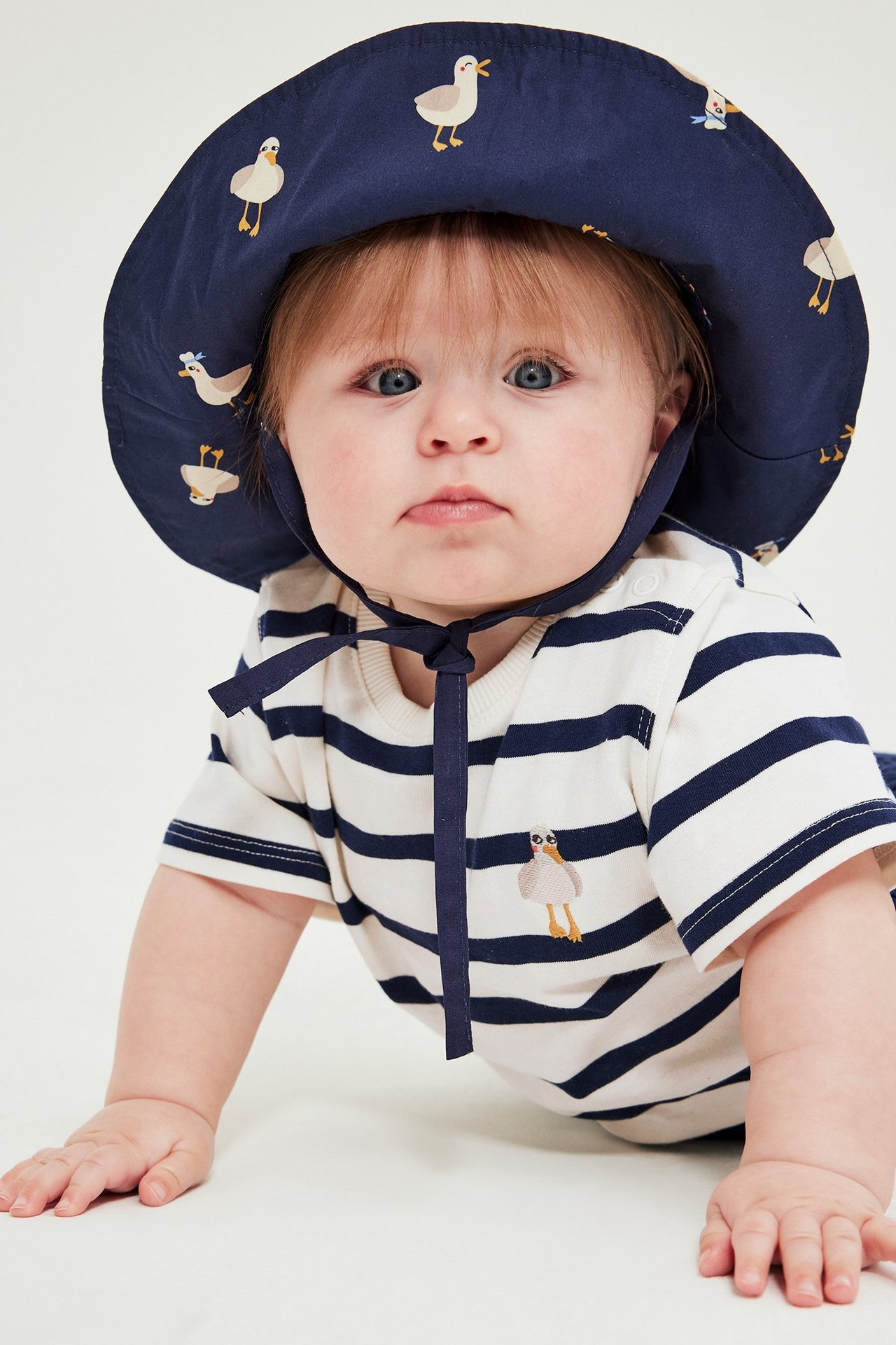 Forfra: baby i stripete bomullsbody med broderi og m mørkblå hatt med måketrykk.