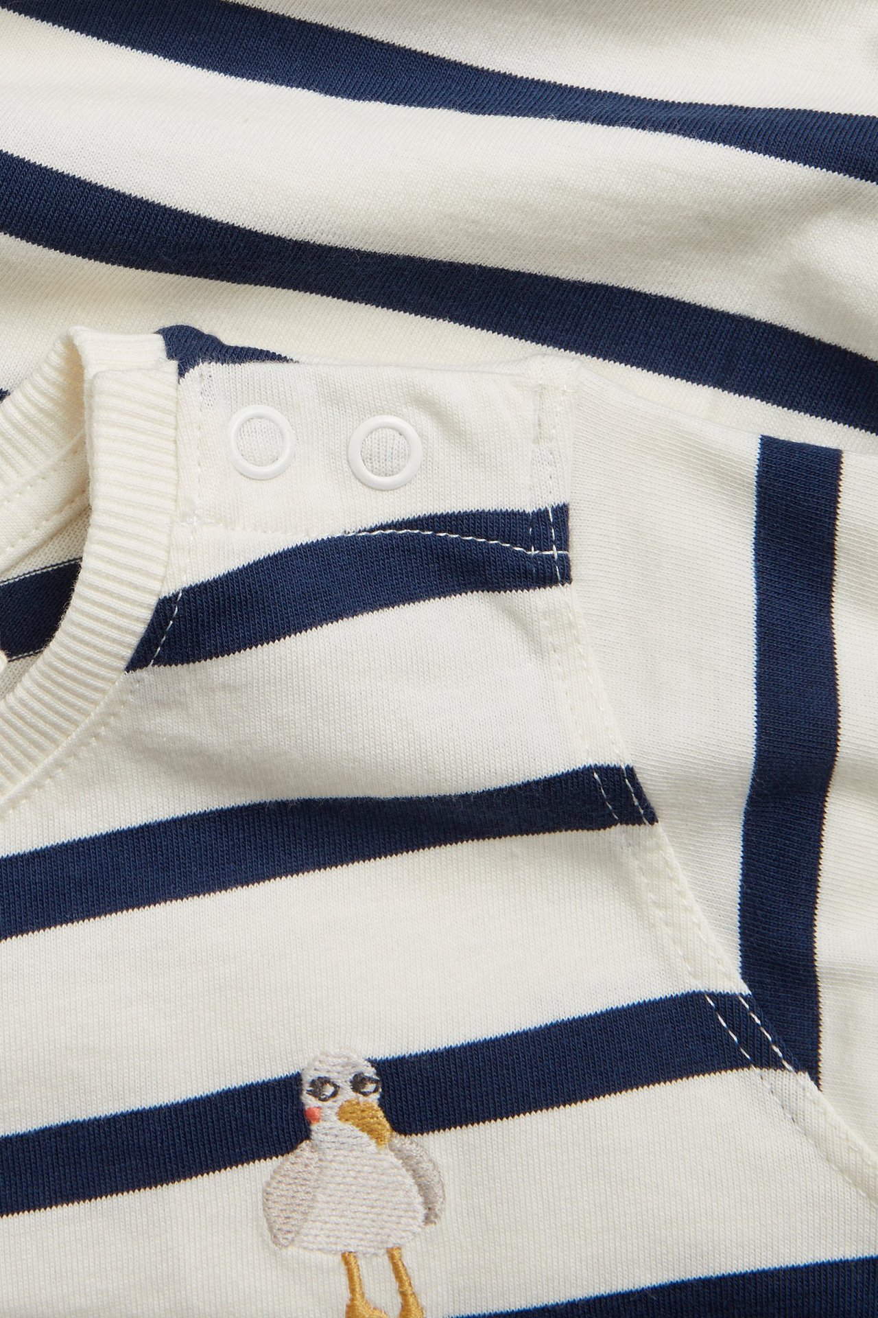 Nærbilde av en stripete babybody i kremhvitt og marineblått med skulderknapper og en måkebroderi.
