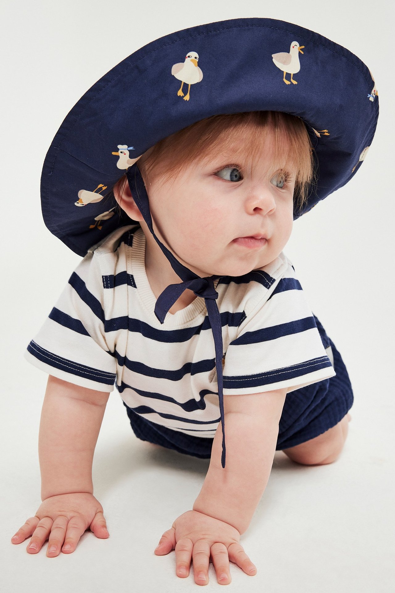 Forfra: baby i hvit og marineblå stripet bomullsbody, marineblå lue med måkemønster og shorts.