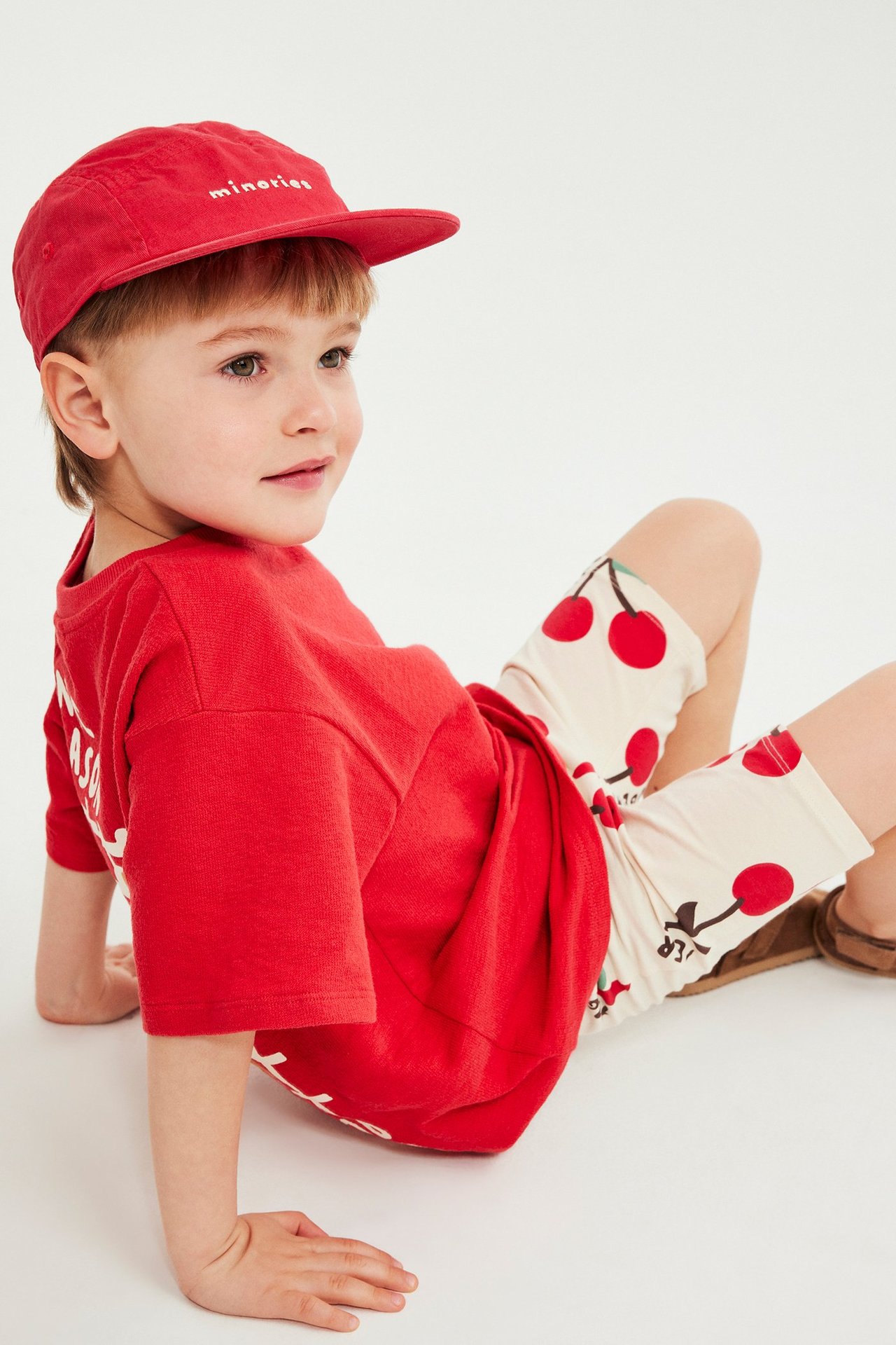 Widok z profilu dziecka w beżowych kolarkach w wiśnie, czerwonym T-shircie i czerwonej czapce.