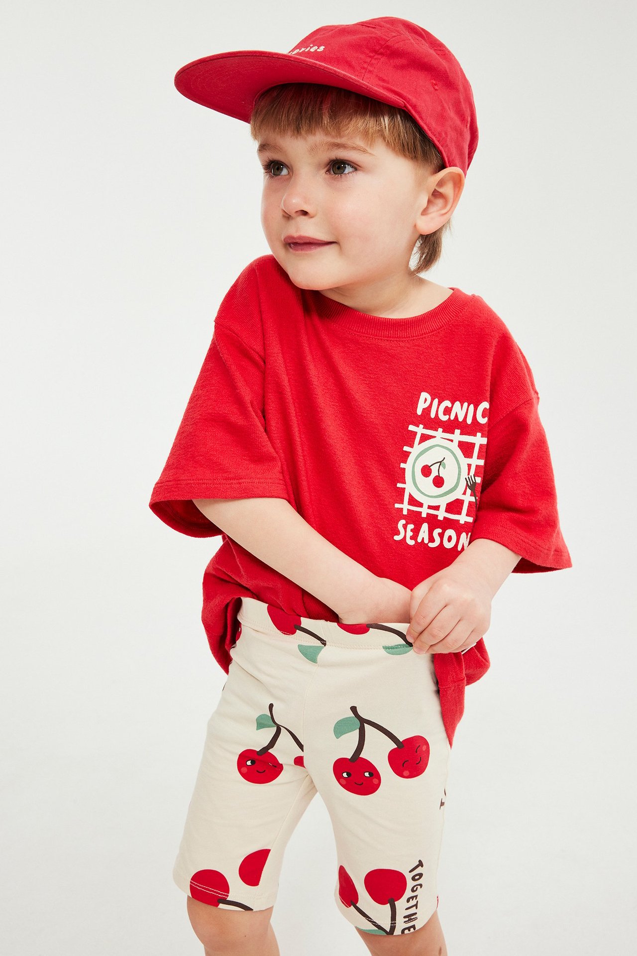 Frontvy av ett barn i beiga cykelbyxor med körsbärsmönster, en röd t-shirt och en röd keps.