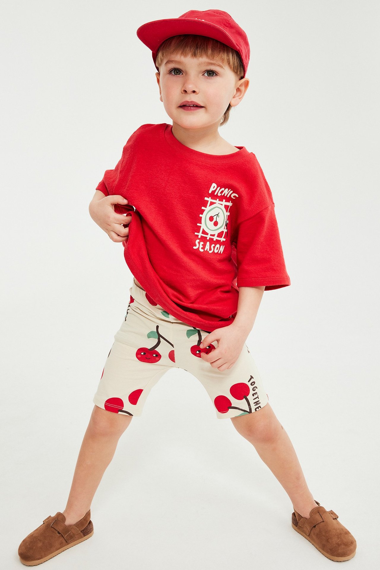 Frontvy av ett barn i beiga cykelbyxor med körsbärsmönster, en röd t-shirt, en röd keps och bruna skor.