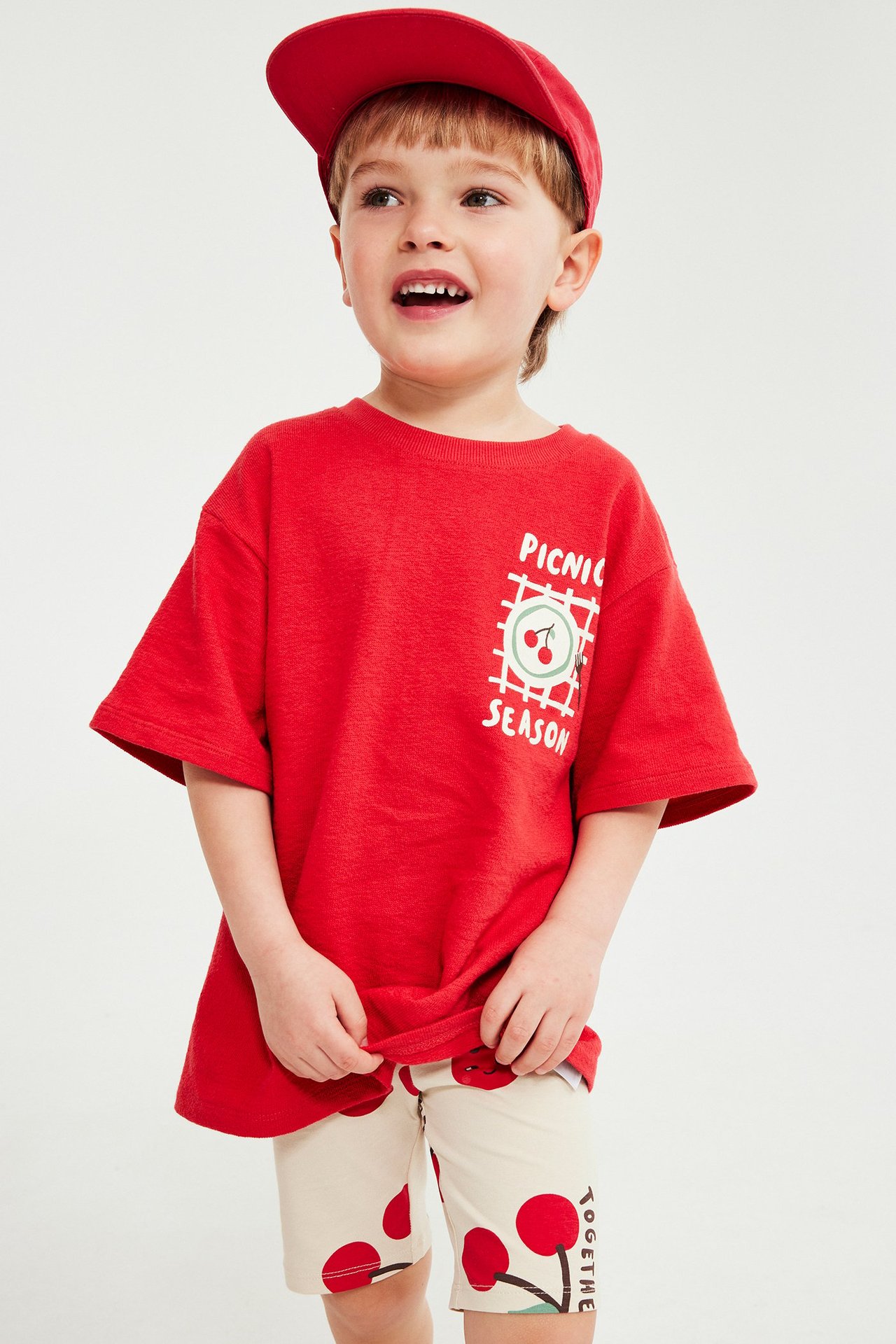 Widok z przodu dziecka w beżowych kolarkach z wiśniami, czerwonej koszulce i czerwonej czapce.