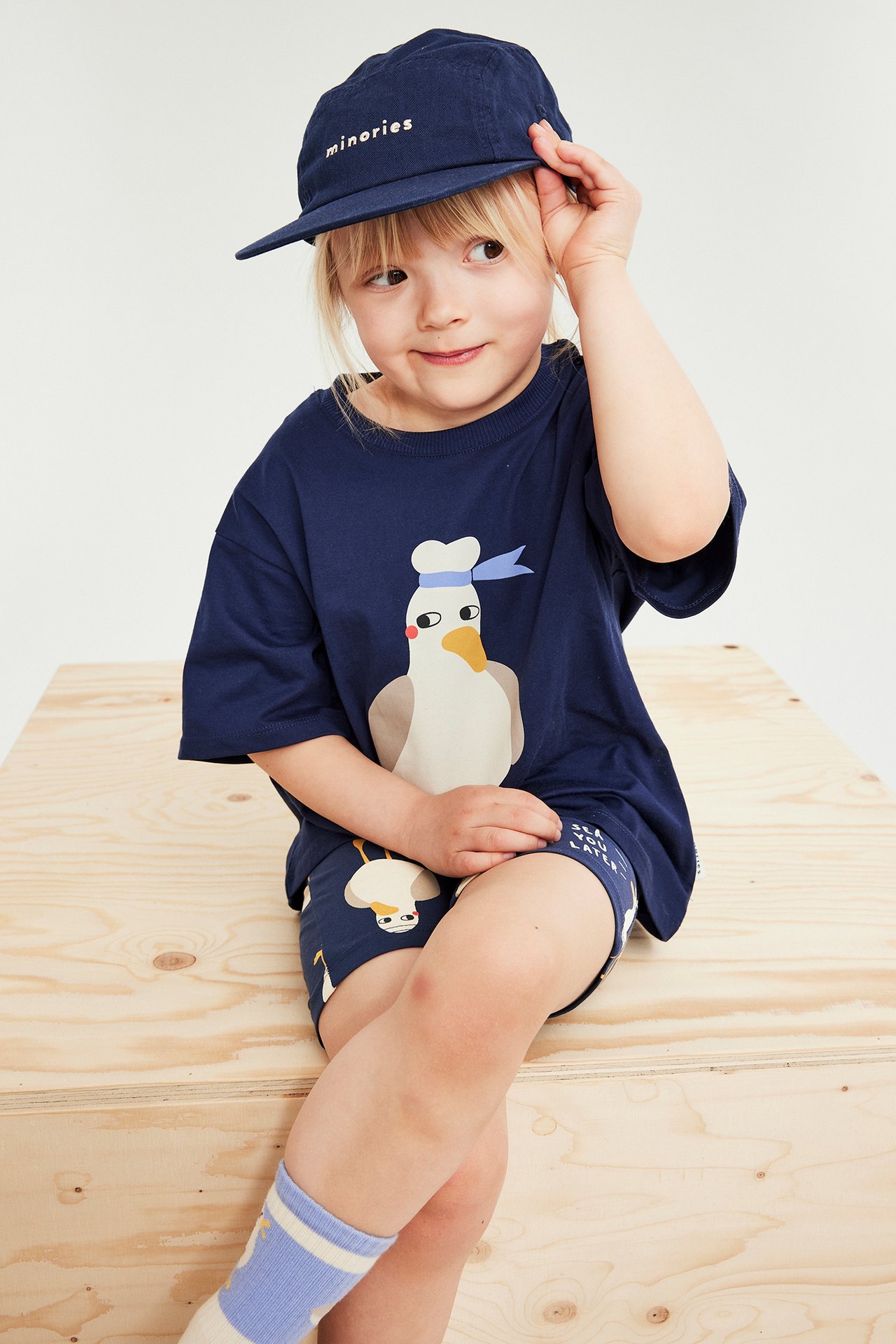 Frontvy av ett barn i blå mönstrade cykelbyxor, en matchande t-shirt, marinblå keps och randiga strumpor.