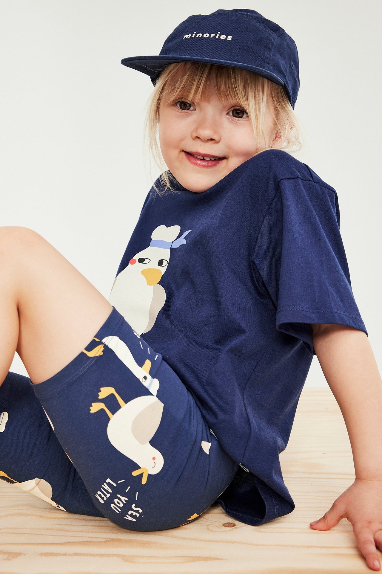 Framifrån, ett barn i blå cykelbyxor med anktryck, en mörkblå t-shirt med mås och en mörkblå keps.