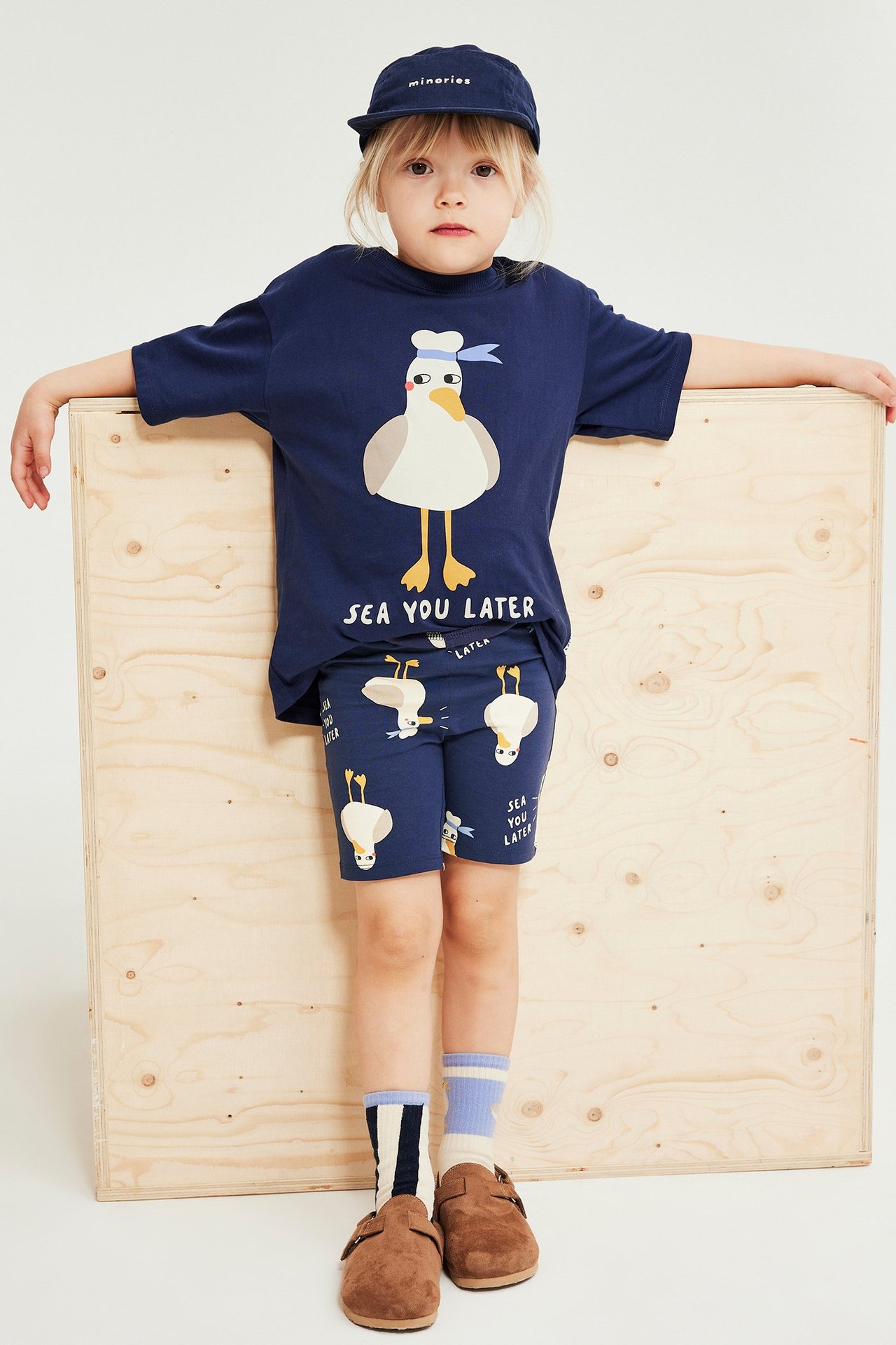 Framifrån: ett barn bär blå mönstrade cykelbyxor med ankor, marinblå tröja, keps, randiga strumpor, bruna träskor.