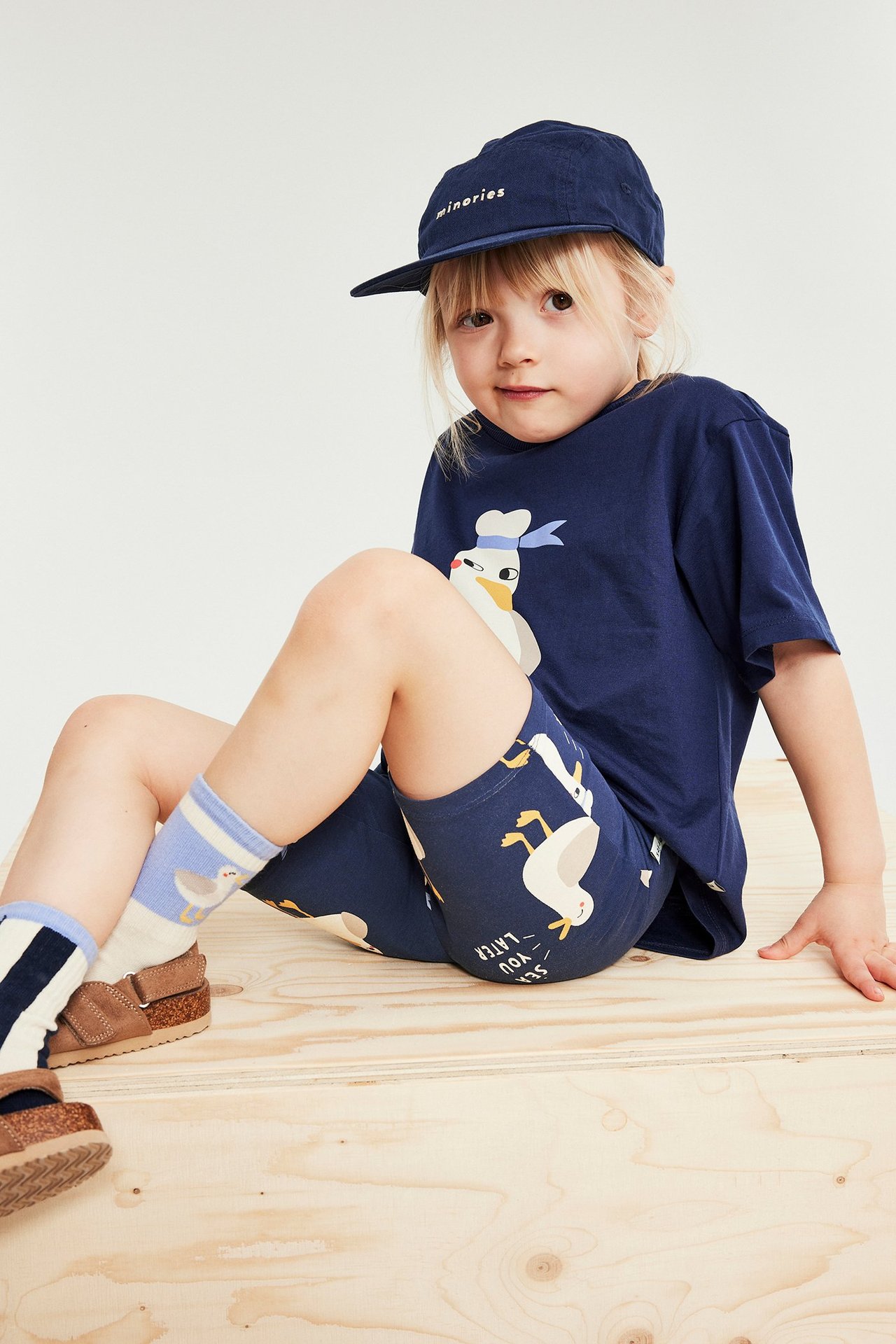 Framifrån: barn bär blå cykelbyxor med ank-mönster, marinblå t-shirt, keps, randiga strumpor och bruna sandaler.