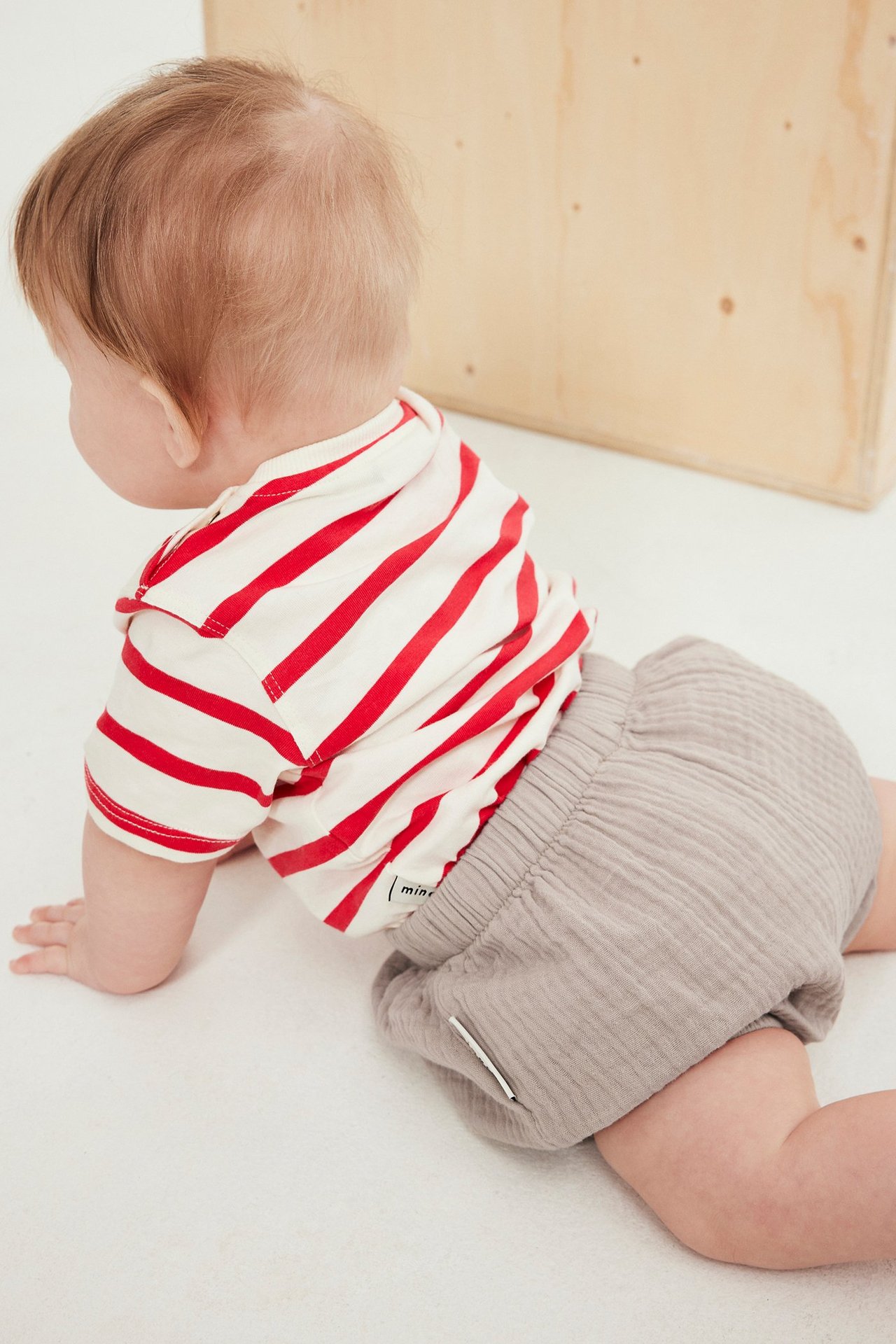 Ryömivä vauva takaa päin, yllään beiget kudotut shortsit ja raidallinen t-paita.