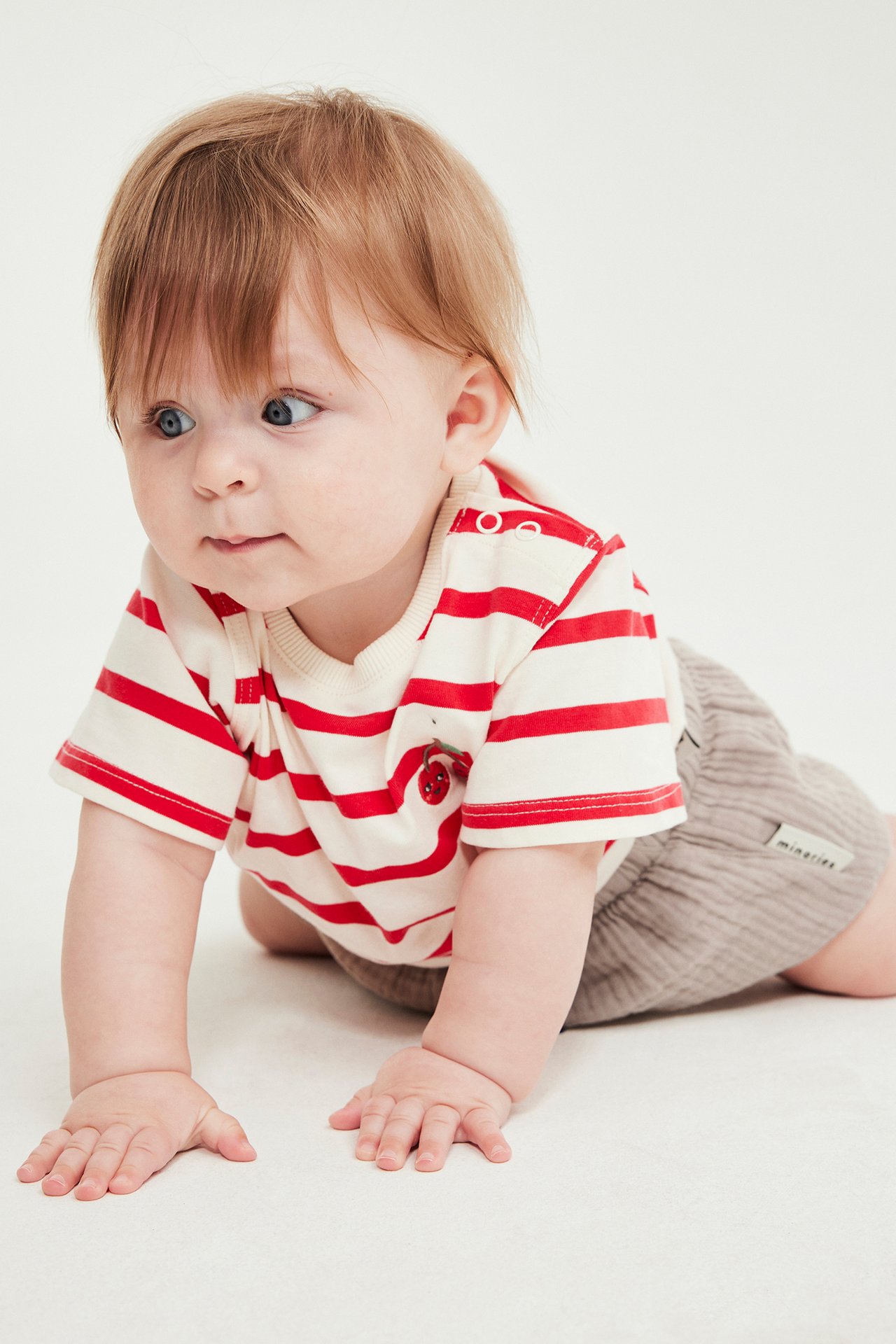 Från sidan, baby som kryper i beige vävda shorts med resår i midjan och en randig t-shirt.