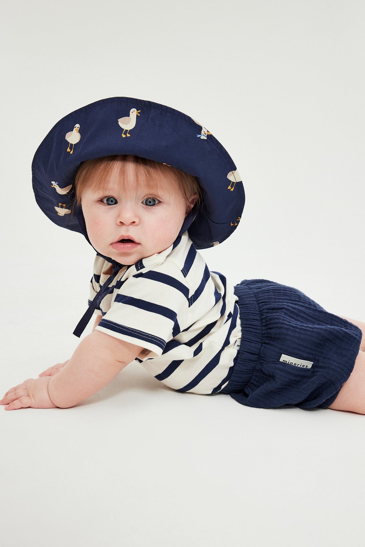 Vauva edestä: siniset kudotut shortsit joustavalla vyötäröllä, raidallinen t-paita ja ankka-kuvioinen aurinkohattu.