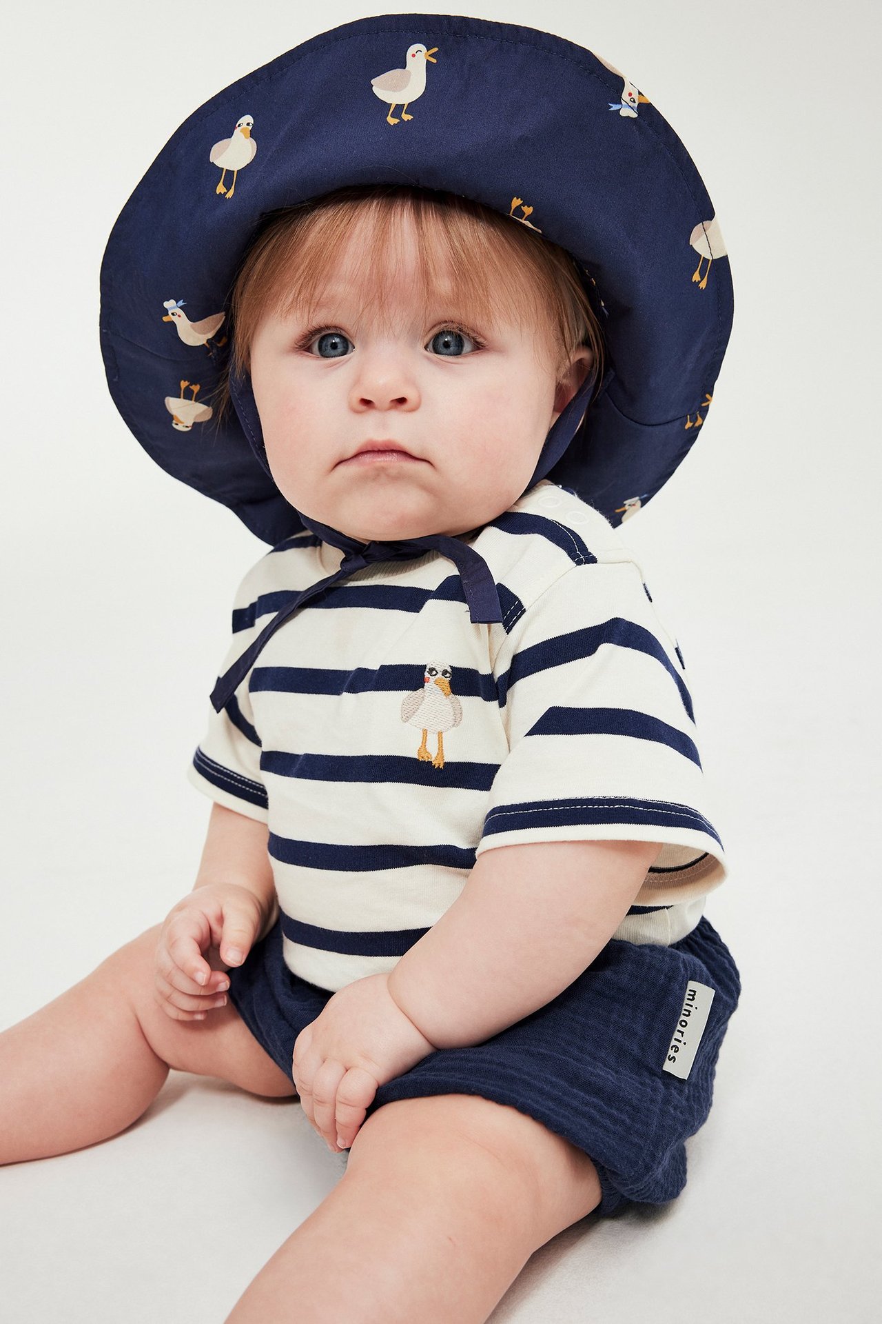 Widok z przodu: niemowlę w niebieskich tkanych szortach, pasiastym podkoszulku i granatowym kapeluszu z ptasim wzorem.