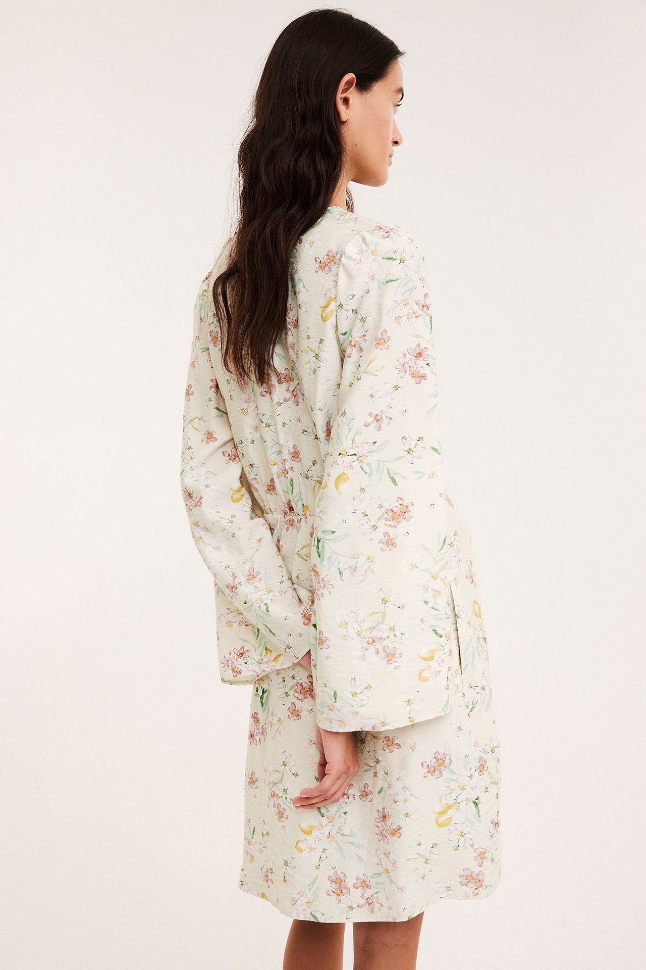 Sidevisning av en kvinne i en lys beige blomstret A-linjet kort kjole med volangermer.