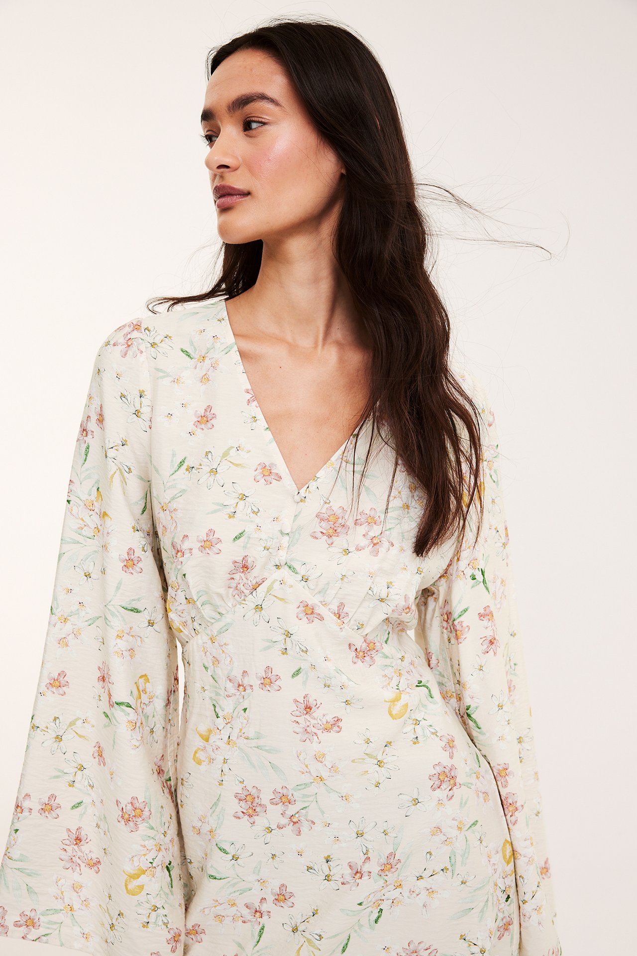 Forfra: Kvinne i lys beige blomstret V-halskjole med knapper foran og vide ermer.