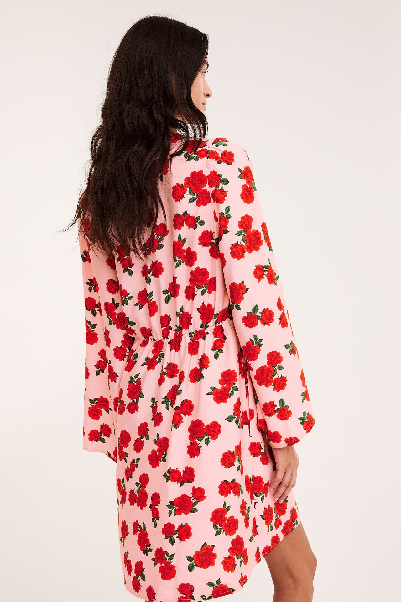 Sida vy av kvinna i kort ljusrosa viskosklänning med rött rosmönster och rynkad midja.