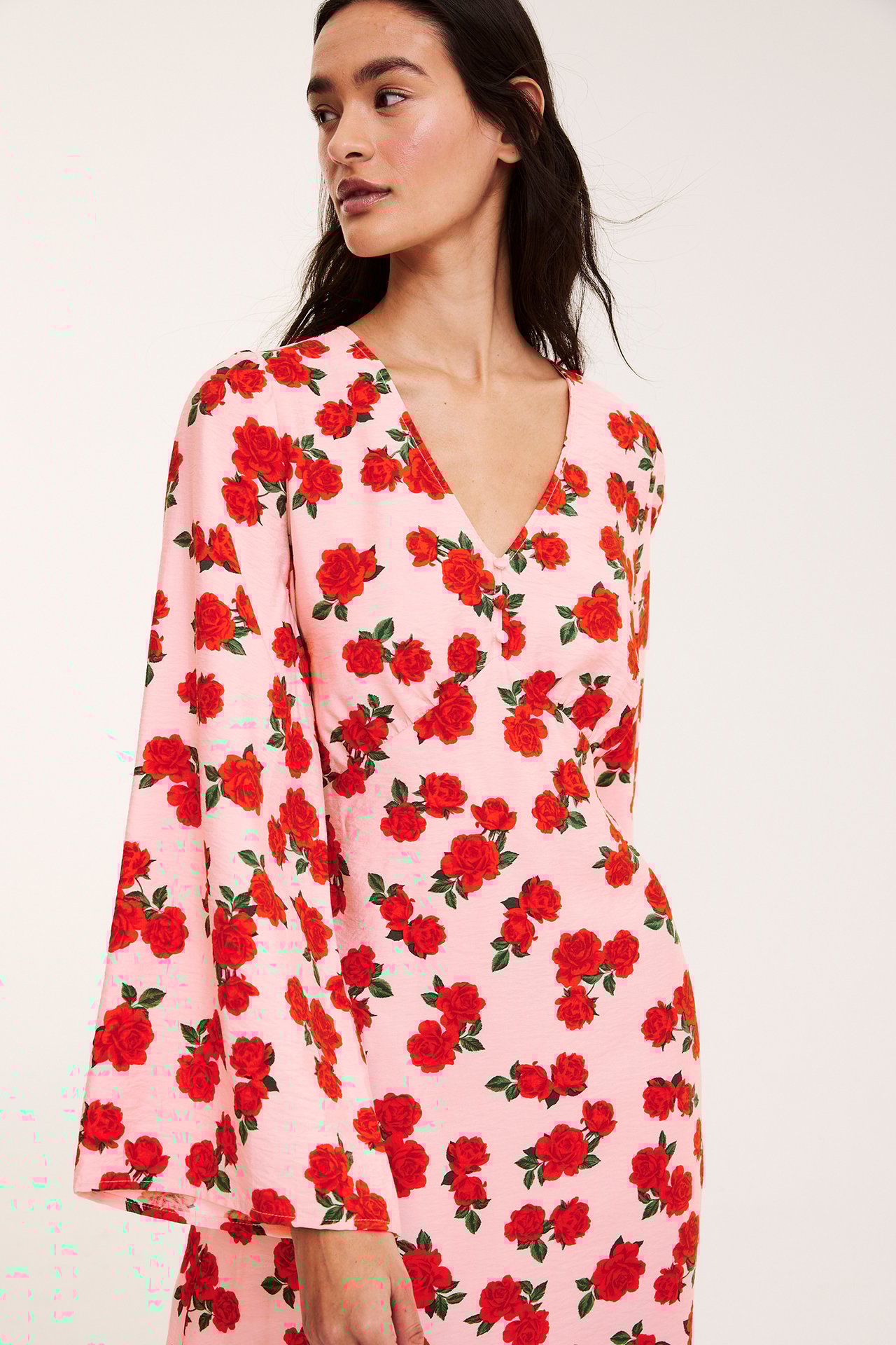 Front-side view av kvinna i ljusrosa A-linjeklänning med röda rosmönster, V-ringning, volangärmar och knappar fram.