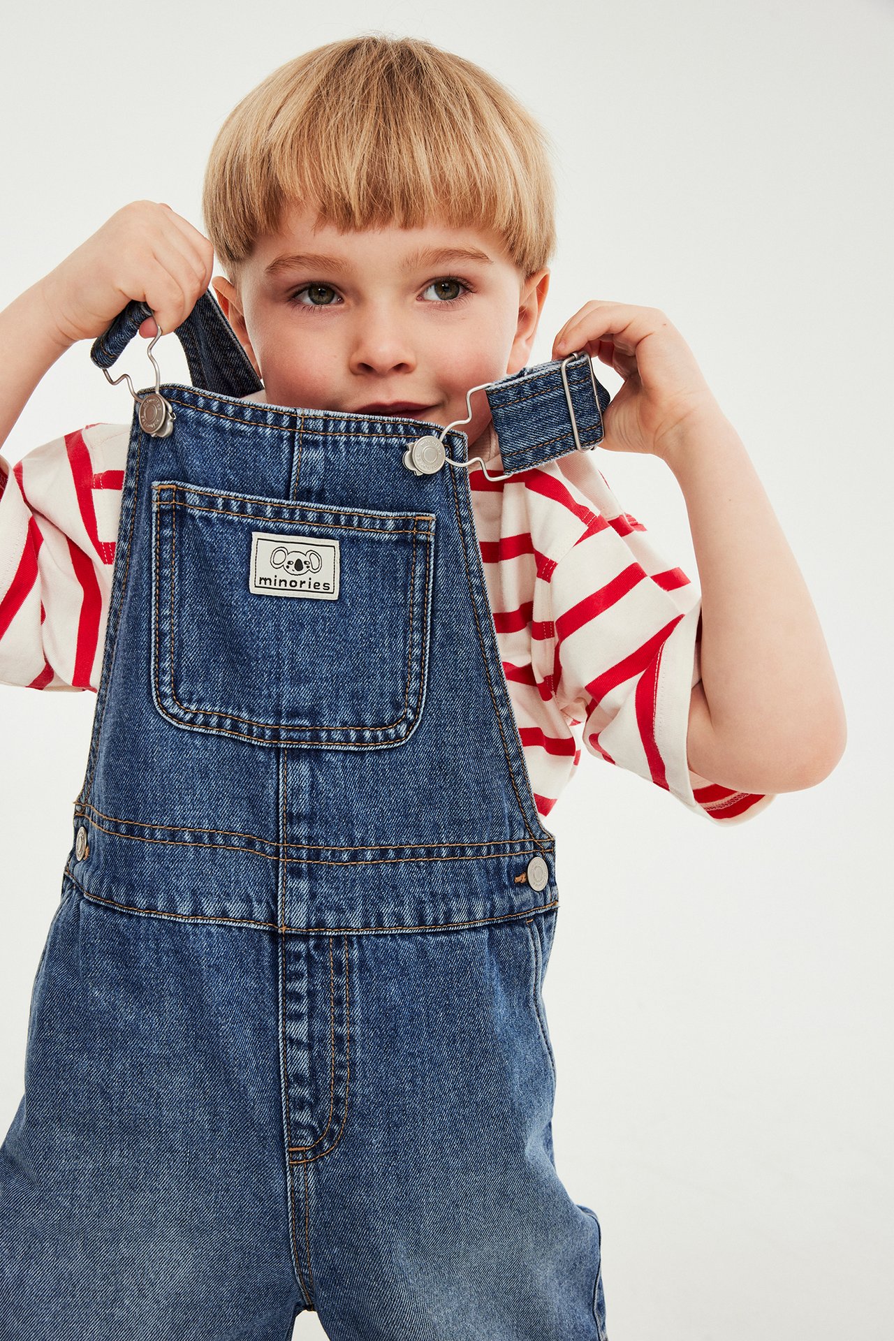 Edestä: Lapsi sinisissä denim-haalarihousuissa valkoisen, punaraitaisen t-paidan päällä, olkaimia pitäen.