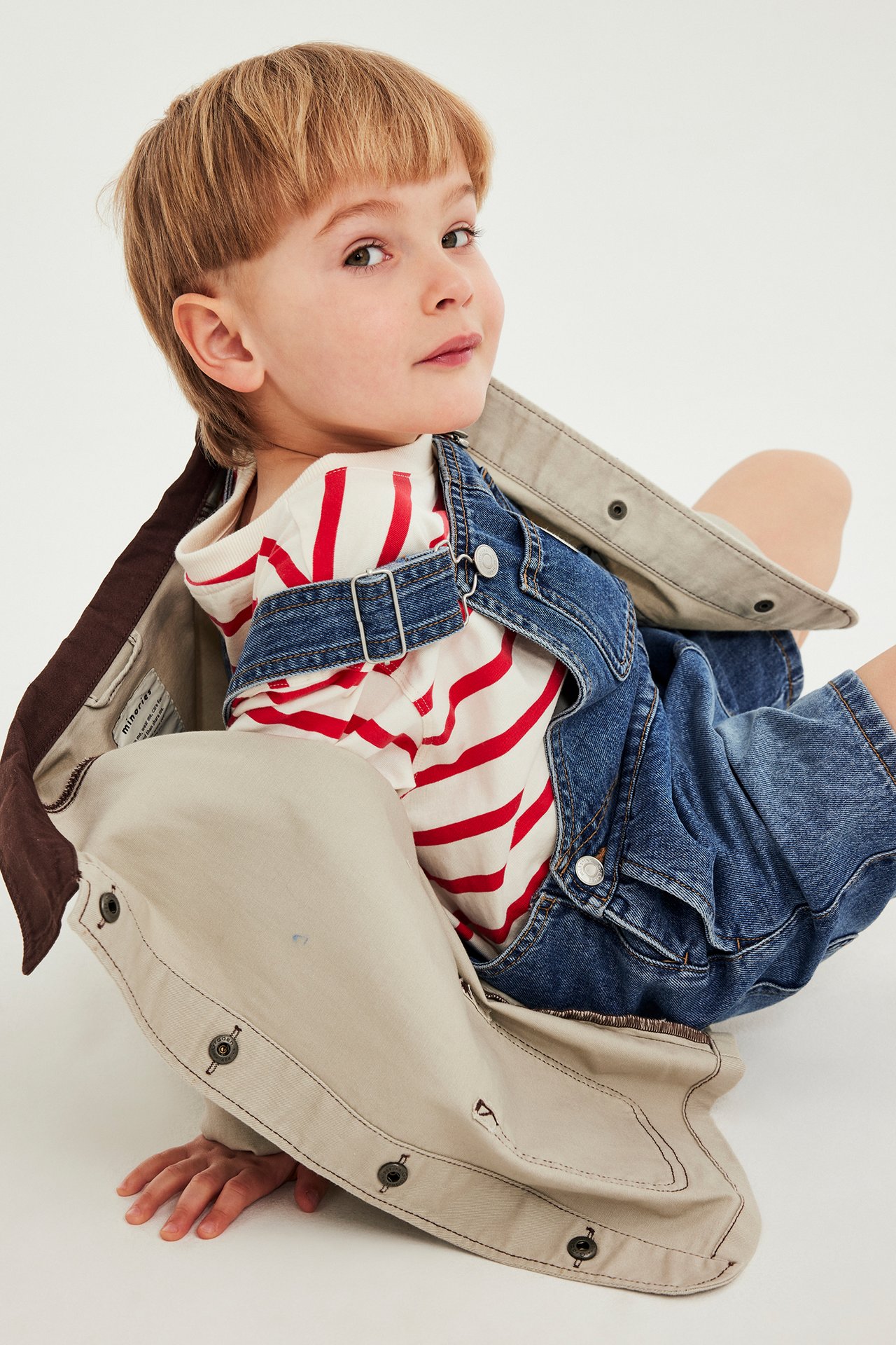 Lapsen osittainen etunäkymä sinisissä denimhaalarihousuissa säädettävin olkaimin, alla punaraitainen t-paita.