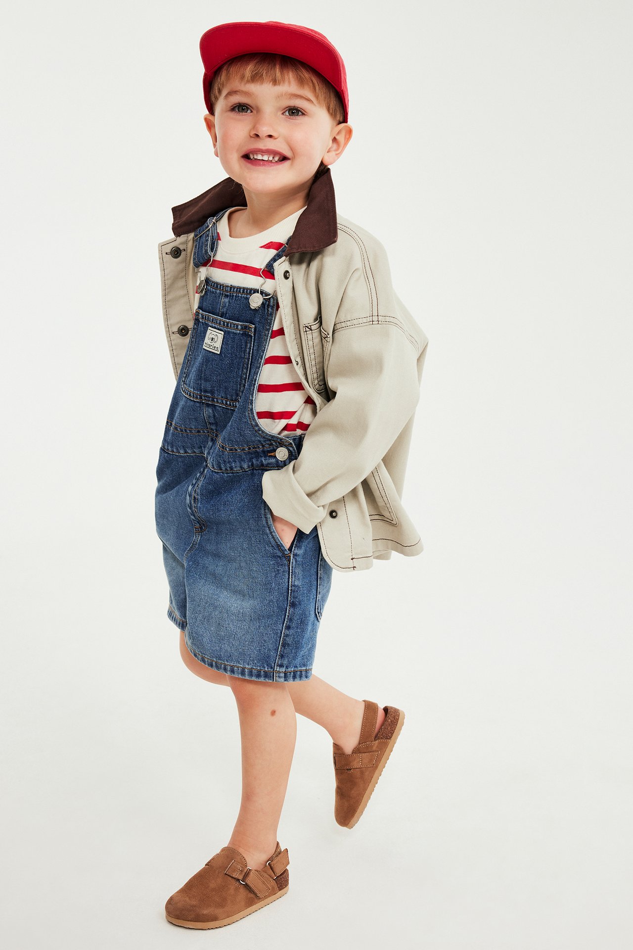 Edestä, lapsi sinisissä denim-haalarihousuissa, raidallinen t-paita, beige takki, punainen lippis, ruskeat kengät.