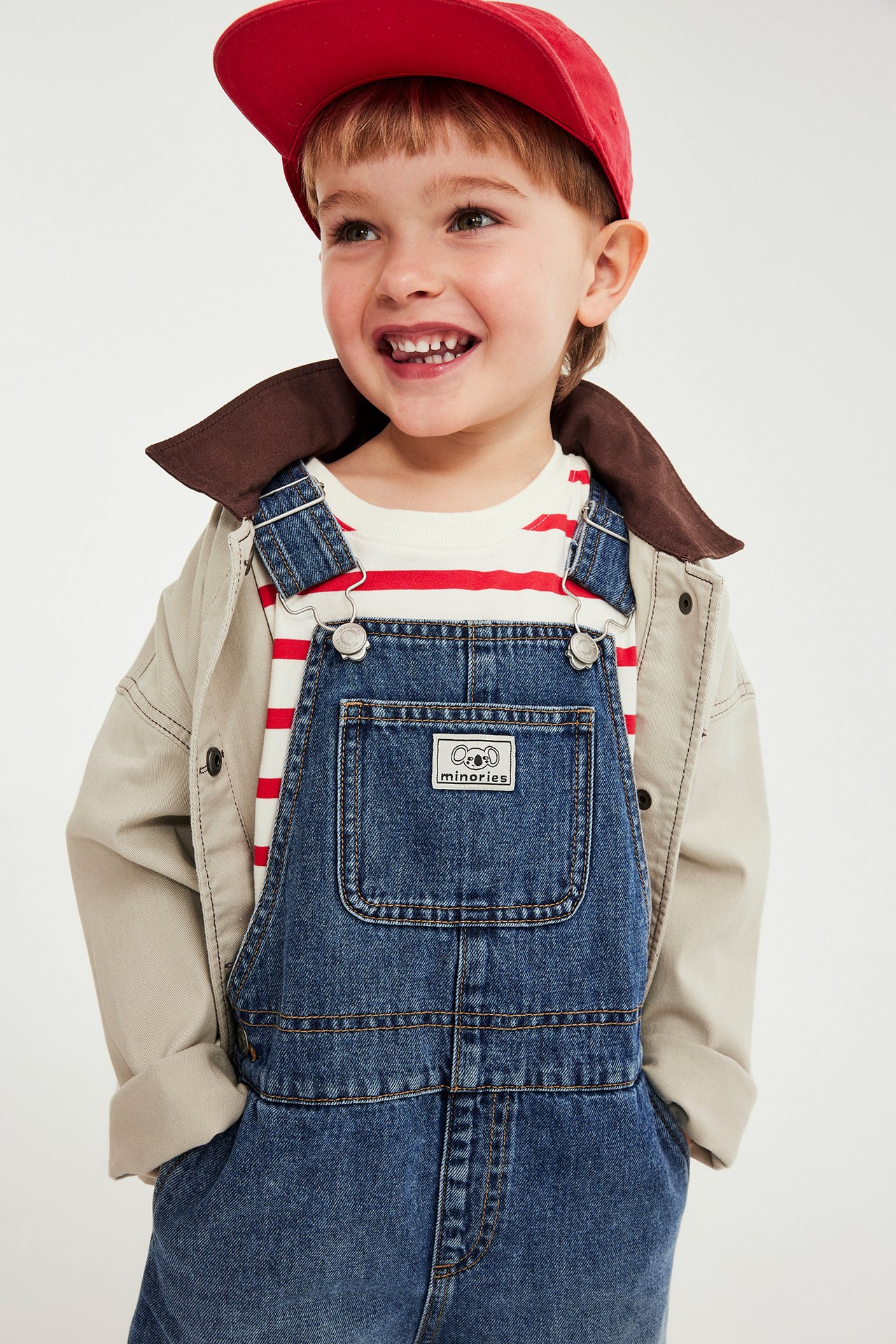 Lapsi sinisissä farkkuhaalareissa, raidallisessa t-paidassa, beigessä takissa ja punaisessa lippiksessä.