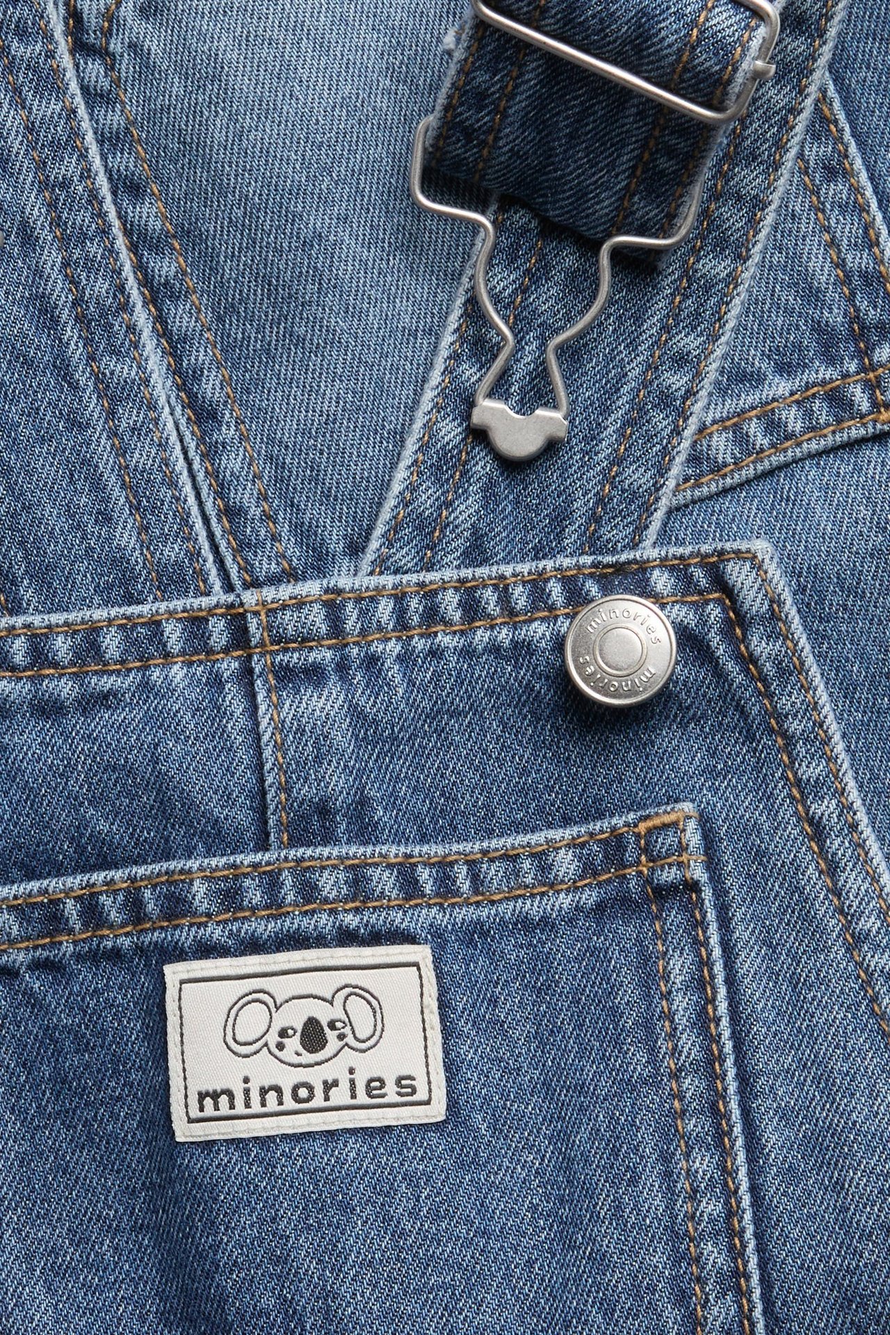 Lähikuva sinisistä farkkuhaalarihousuista säädettävillä metallisoljilla ja Minories-logomerkillä.