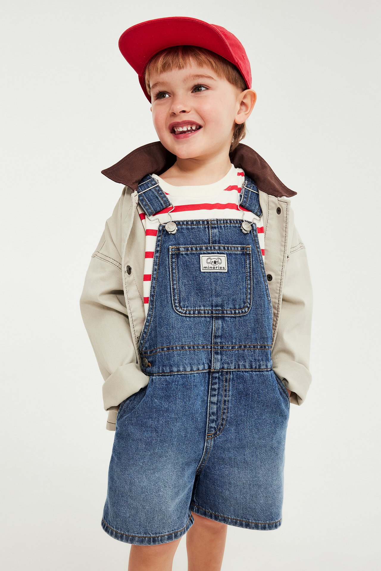 Edestäpäin lapsi yllään siniset denim-haalarihousut, raidallinen t-paita, beige takki ja punainen lippalakki.