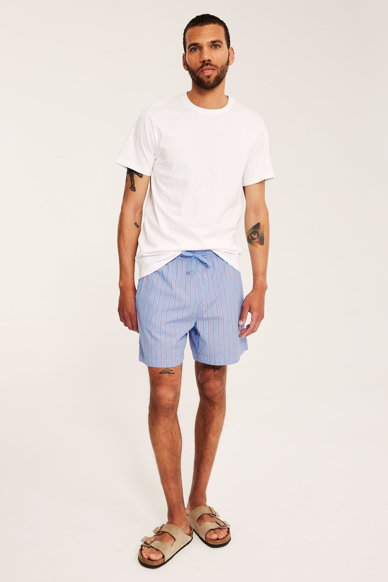 Helkroppsbild framifrån av en man i avslappnade blårandiga bomullsshorts med dragsko, vit t-shirt och beiga sandaler.