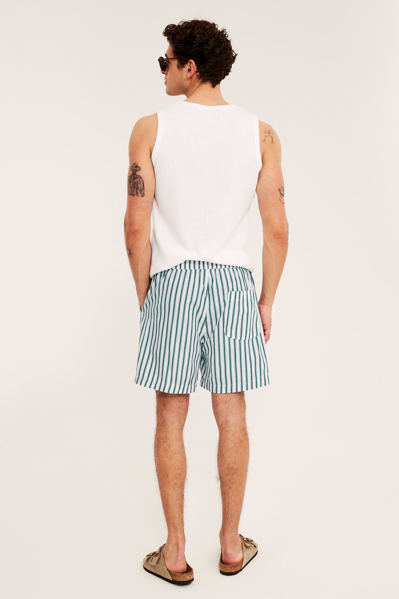 Mann bakfra i hvit- og grønnstripete shorts, en hvit ribbet singlet og brune sandaler.