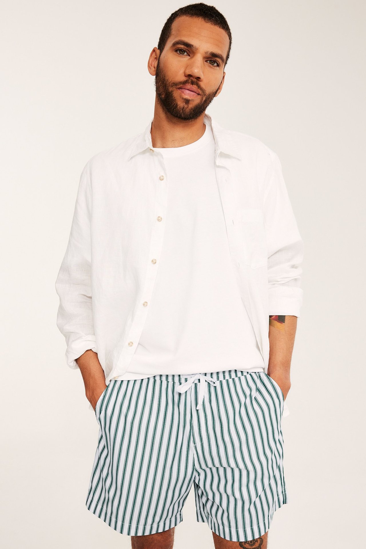 Frontvy: Man i vit- och grönrandiga bomullsshorts, vit t-shirt och öppen vit skjorta.
