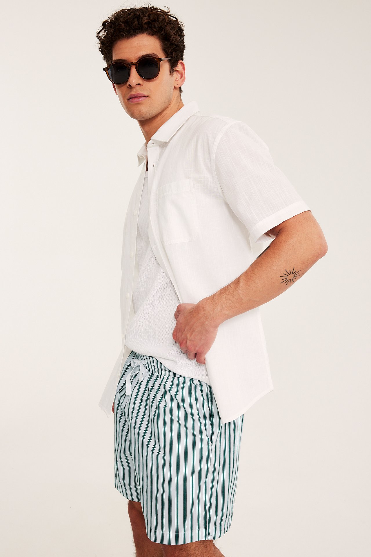 Sida vy: Man i vit-gröna randiga bomullsshorts, vit öppen skjorta, runda bruna solglasögon.