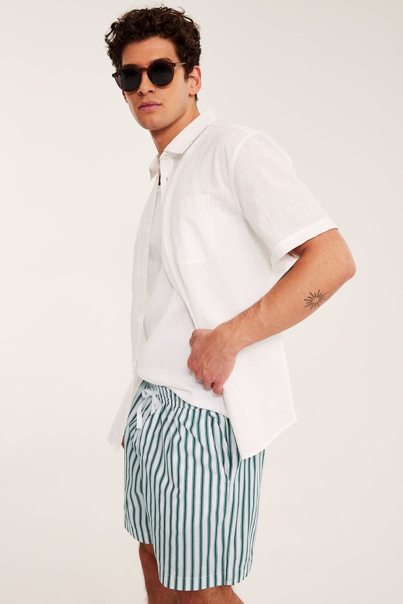 Sida vy: Man i vit-gröna randiga bomullsshorts, vit öppen skjorta, runda bruna solglasögon.